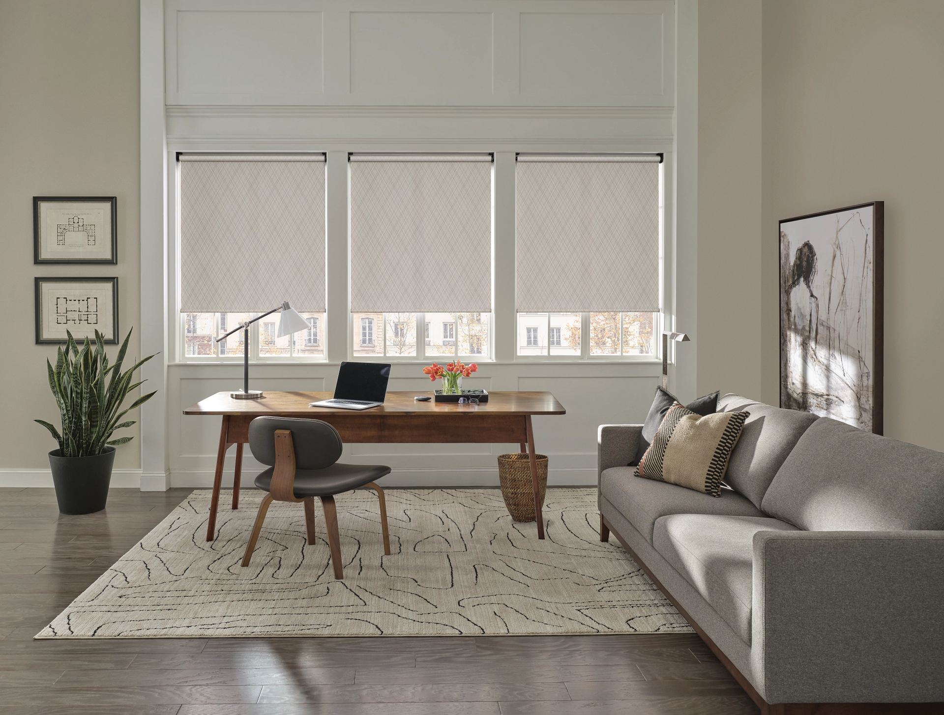 A bright home office with a wooden desk, chair, sofa, and floor-to-ceiling windows featuring neutral roller shades.