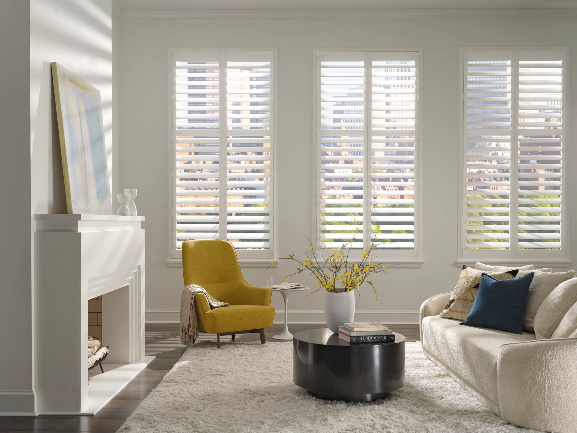 A bright living room with white plantation shutters on three windows, a yellow armchair, and a cream-colored sofa.