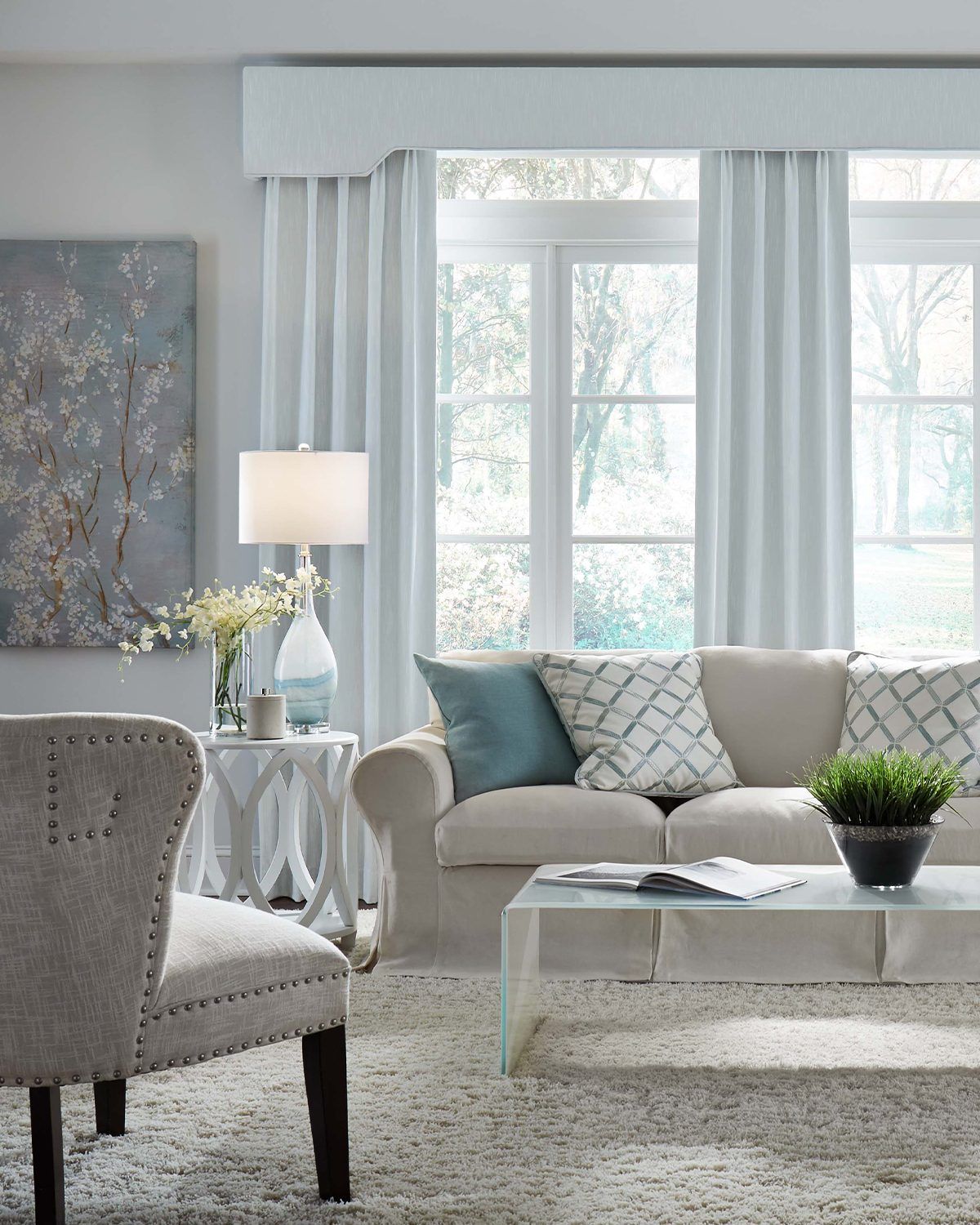 A modern living room with a beige sofa, a patterned accent chair, a coffee table with a plant, and large white curtains.