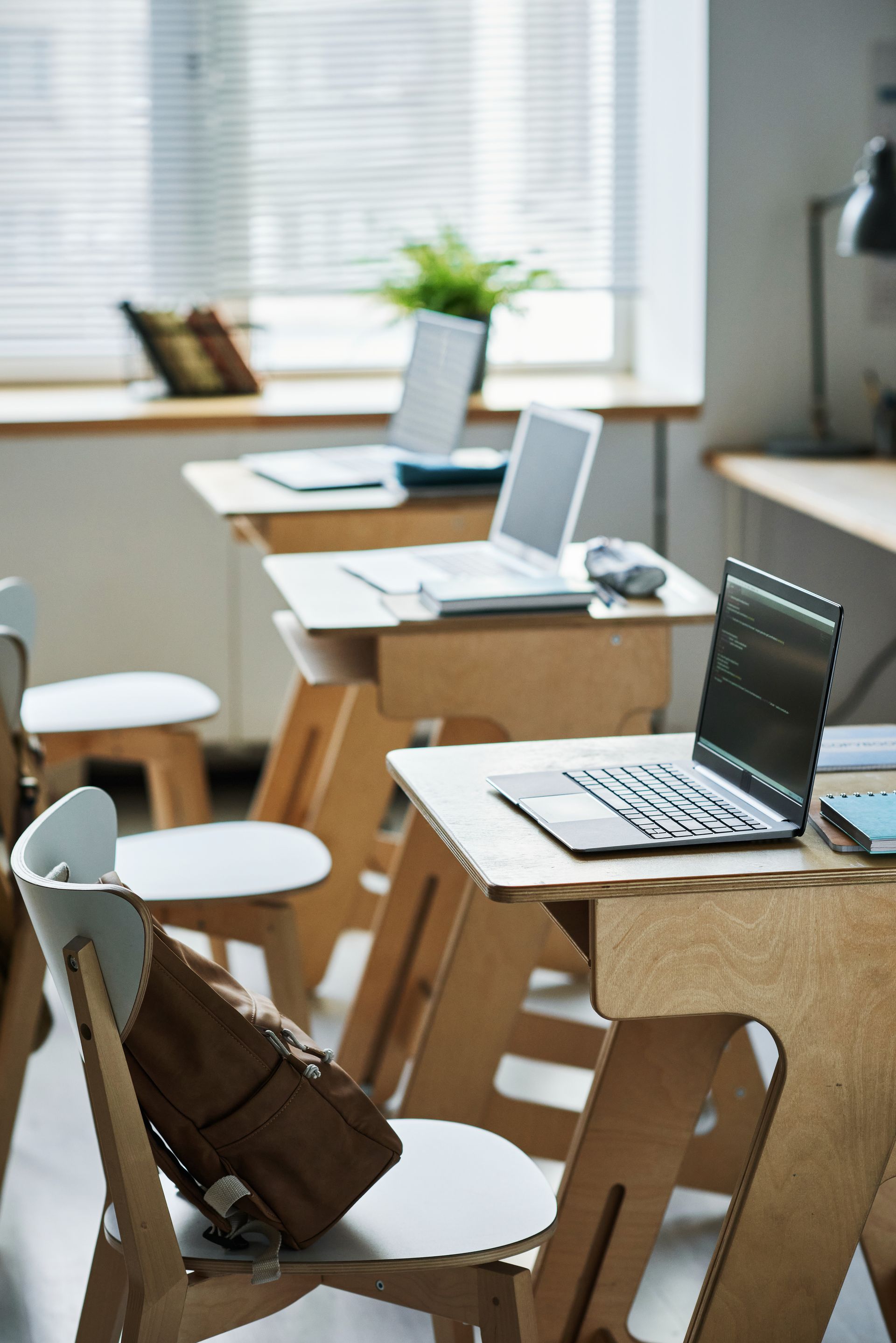 Laptops sit on individual plywood desks arranged in a classroom with a window in the background.