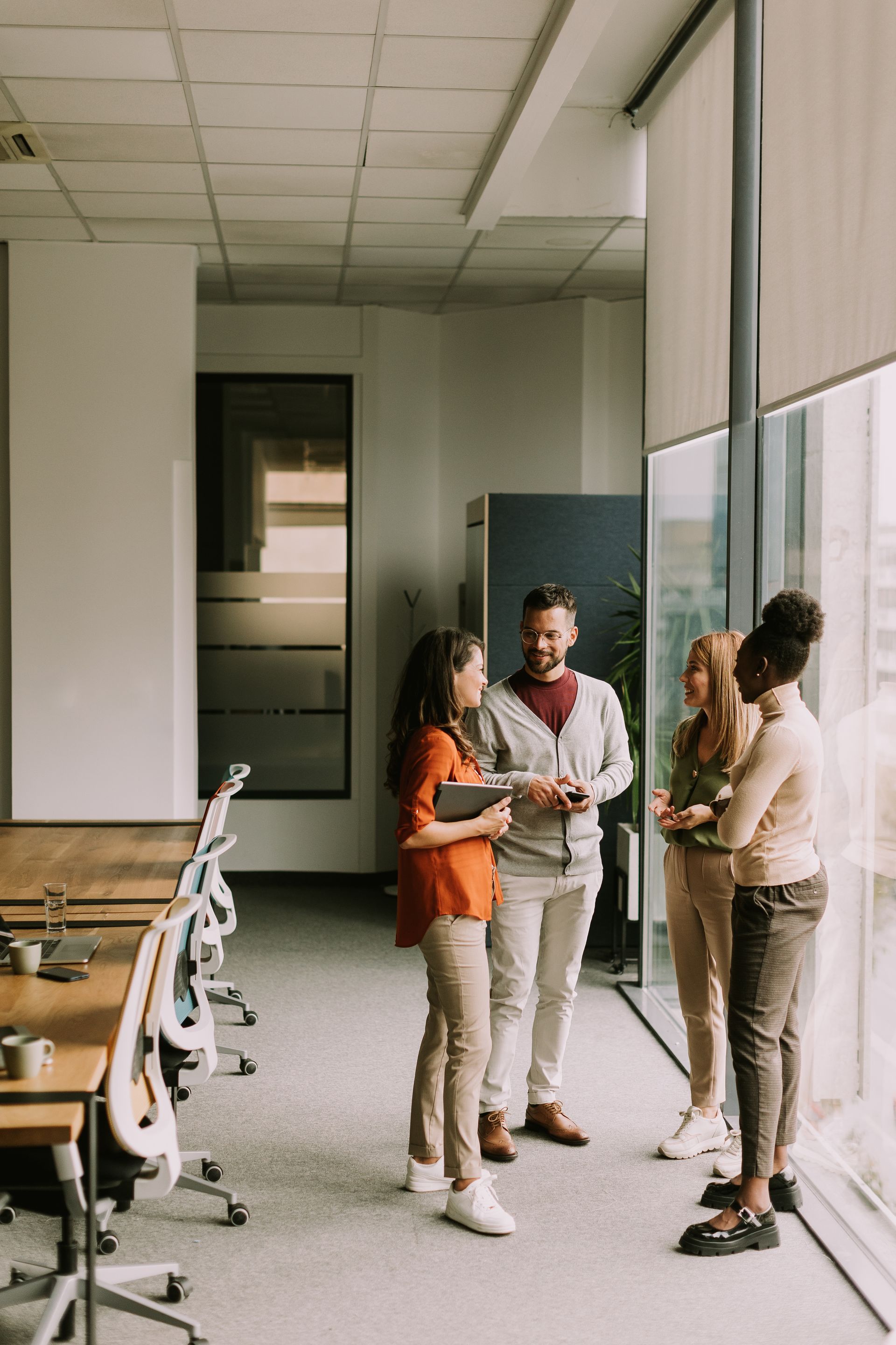 Four colleagues stand in a bright office by a window, talking and smiling while holding tablets.
