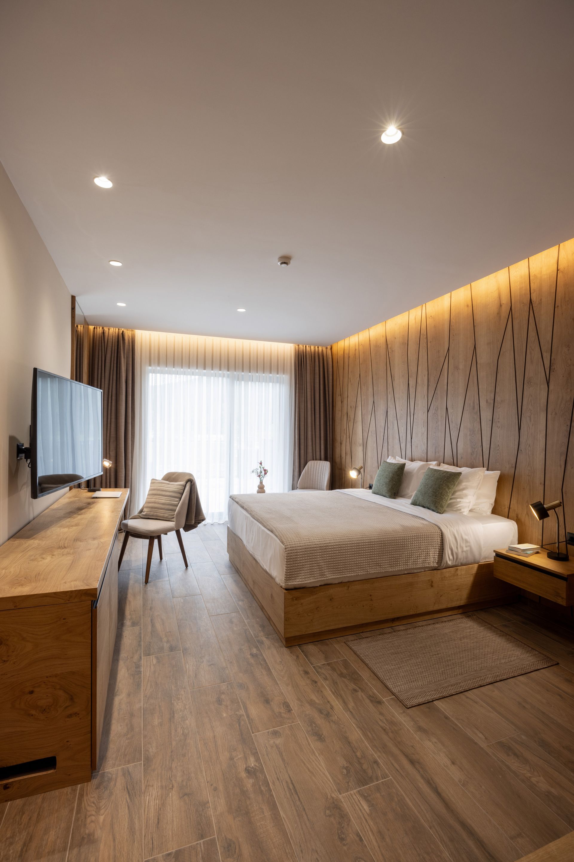 Modern hotel room with a queen bed, wooden headboard, flat-screen TV, arm chair, and floor-to-ceiling window.