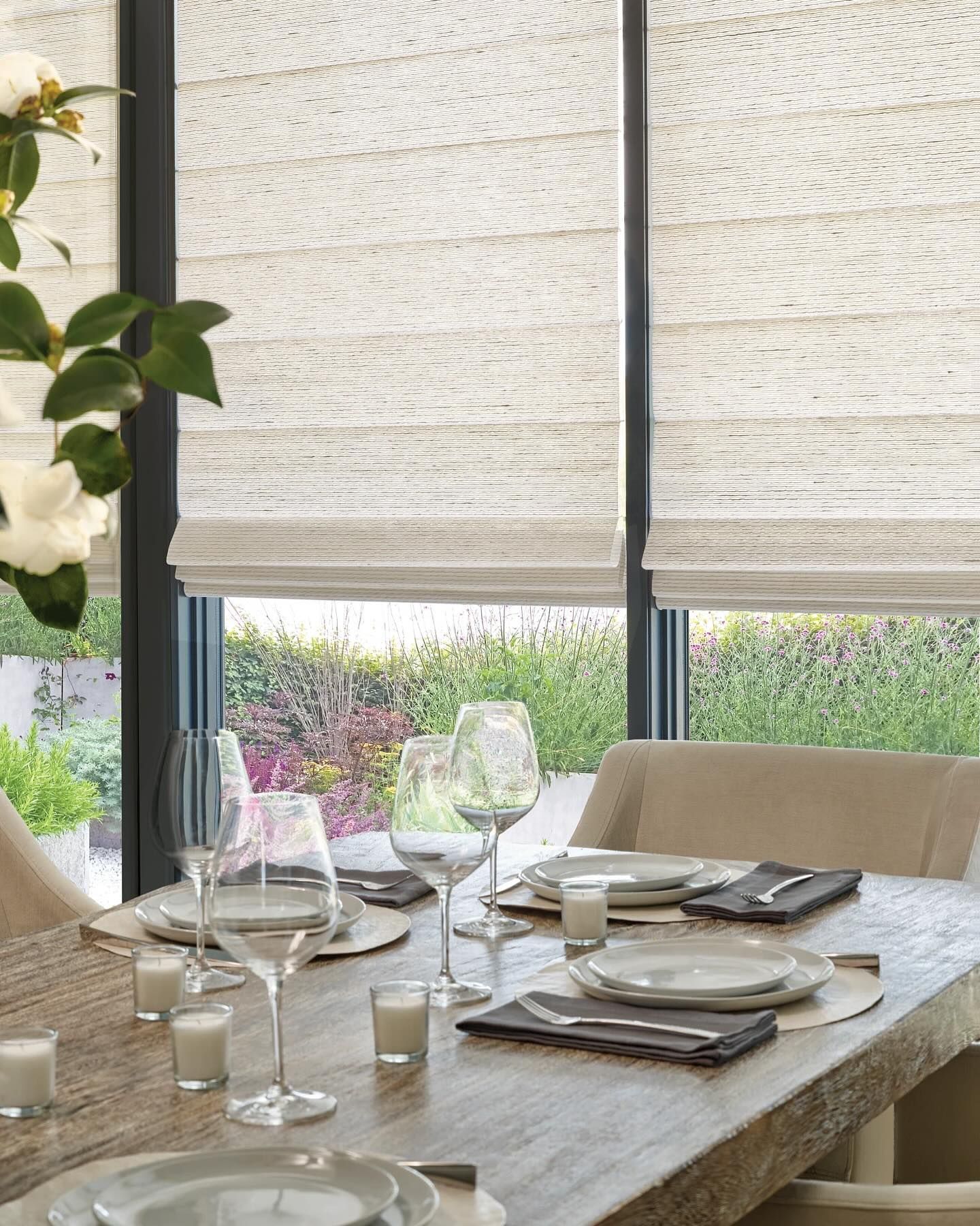 A dining table set for a meal with plates, glassware, and candles, placed in front of large windows with fabric shades.