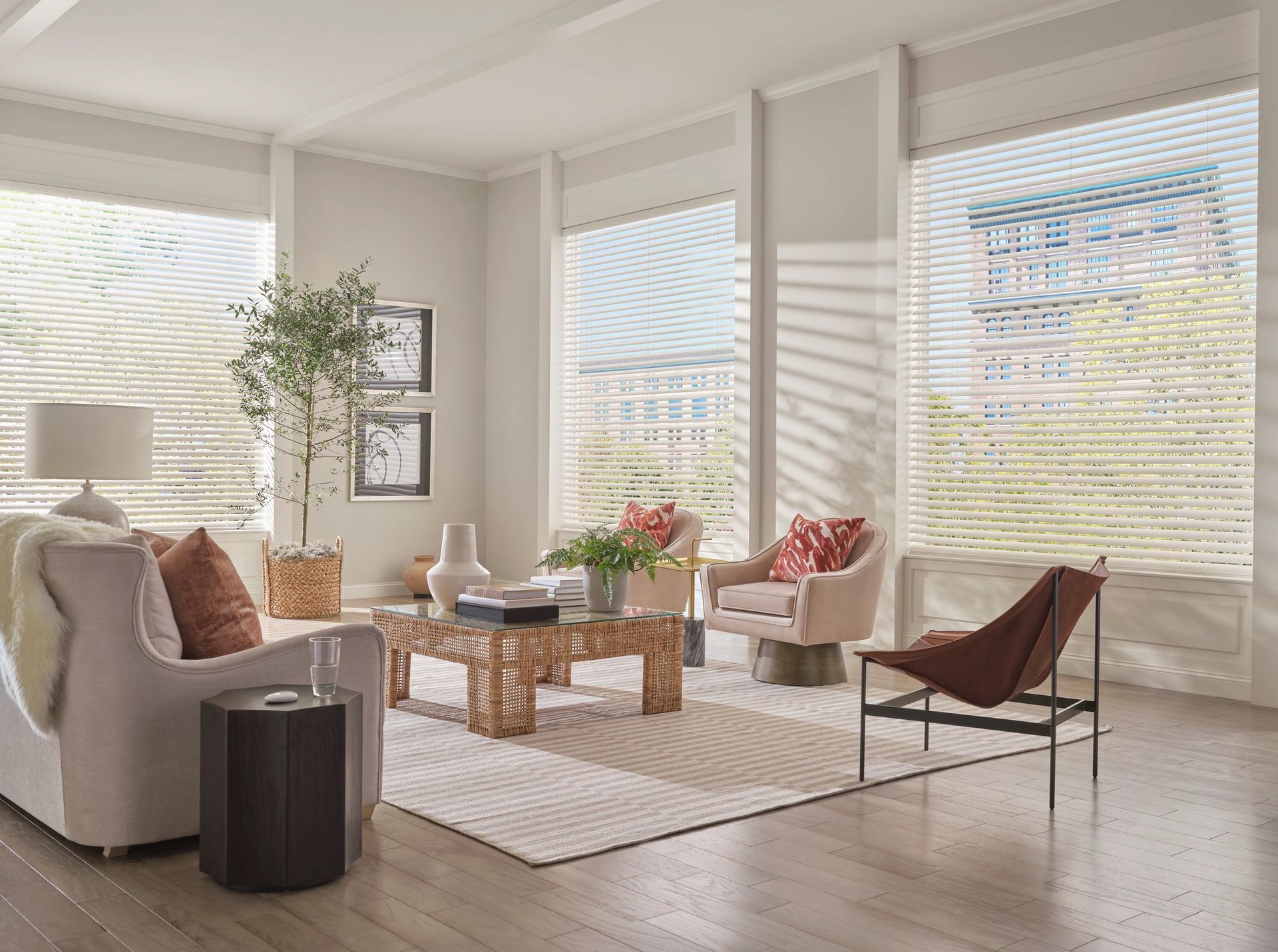 A modern, sunlit living room with light walls, beige furniture, patterned pillows, a wooden coffee table, and large windows.