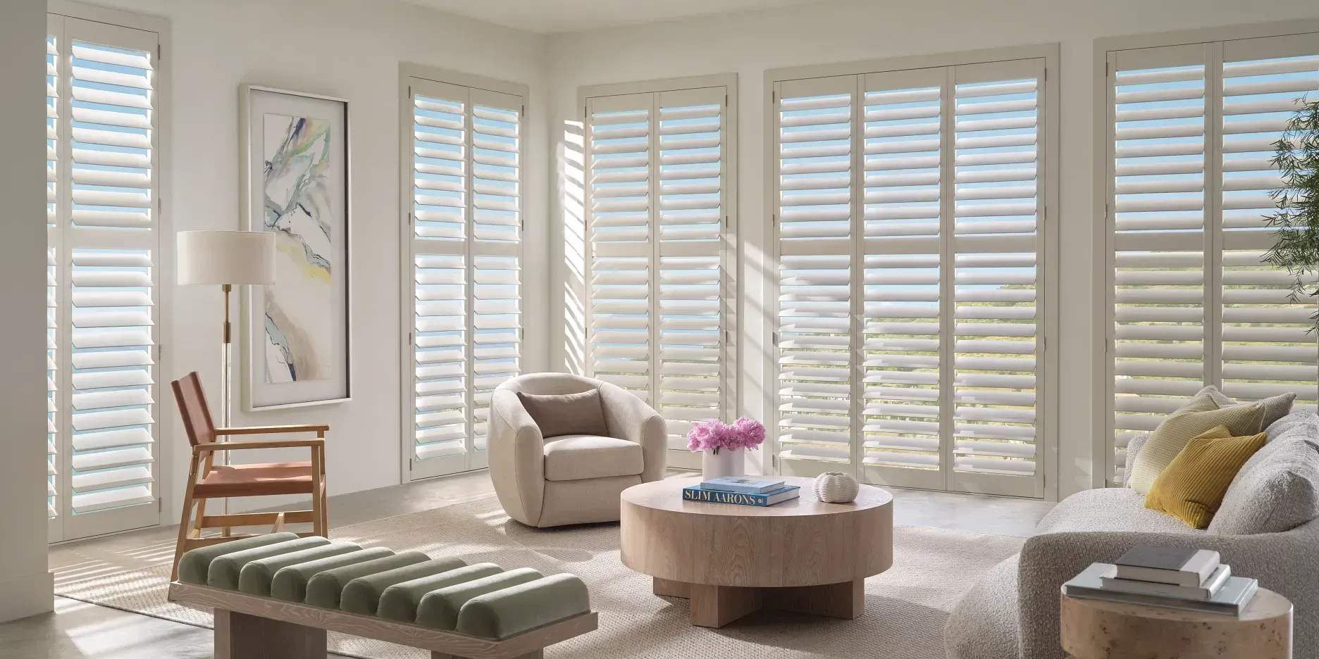 A bright, modern living room with white plantation shutters covering a wall of windows, neutral furniture, and decor.