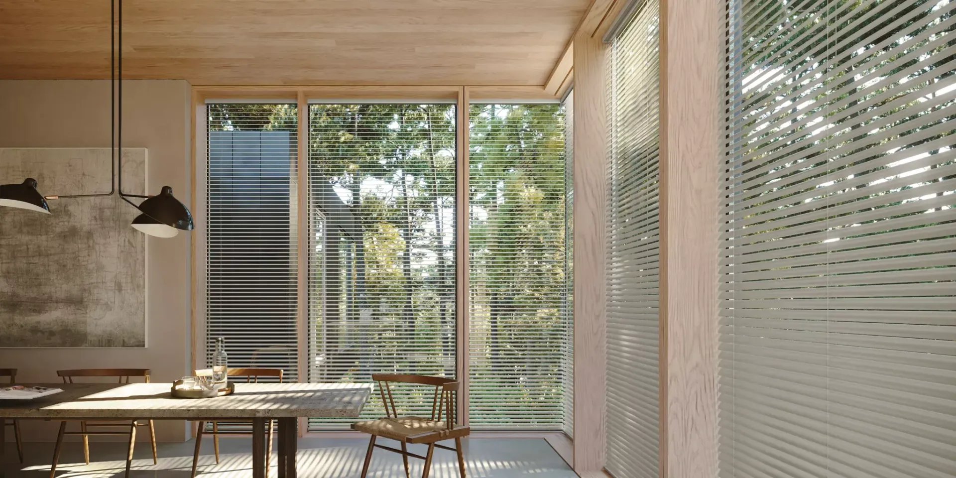 A bright, modern dining room with a wooden table, chairs, and large windows with horizontal blinds overlooking trees.