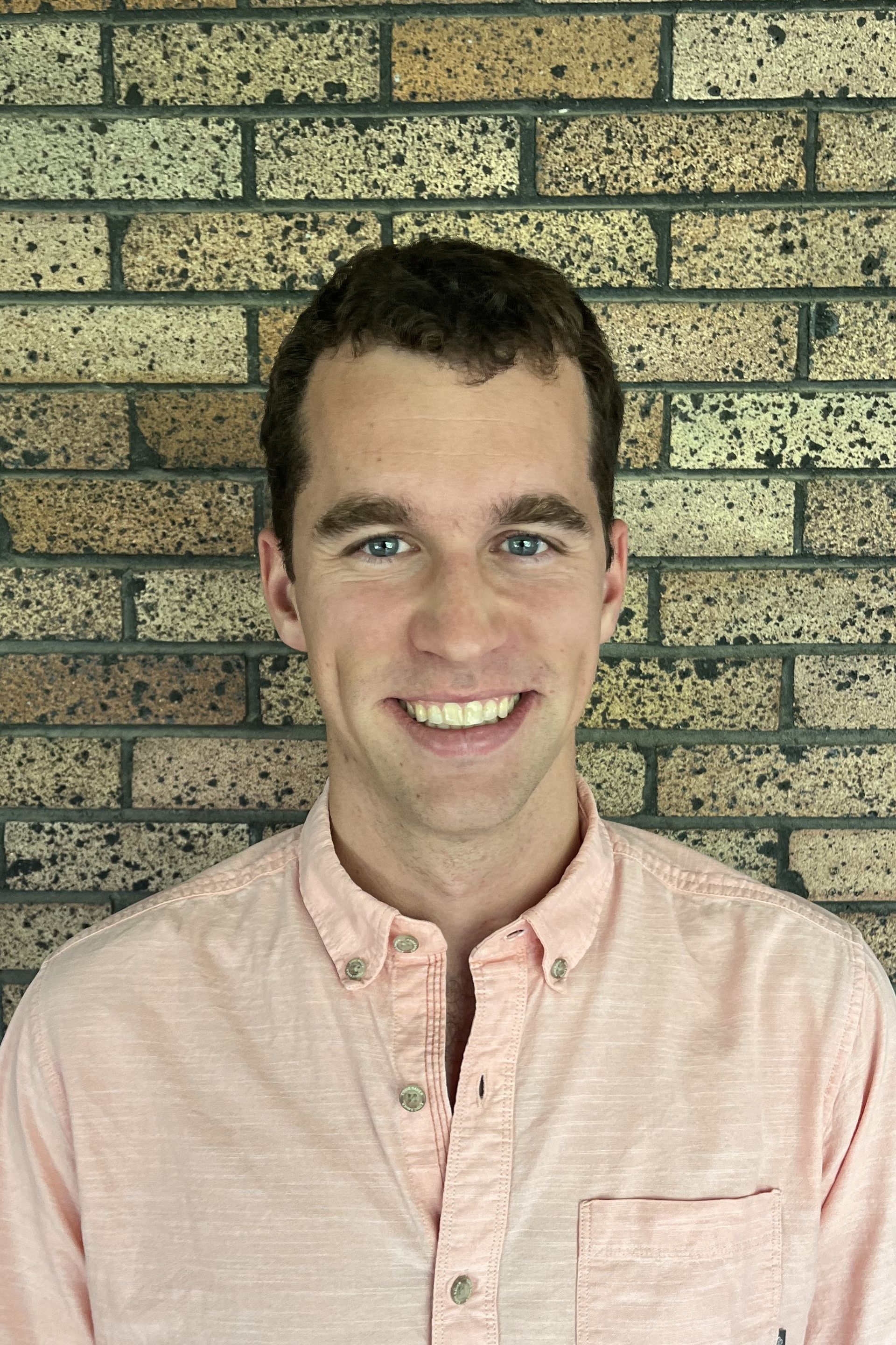 A man in a pink shirt is smiling in front of a brick wall.