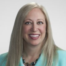 A woman with long blonde hair is smiling and wearing a blue jacket and necklace.