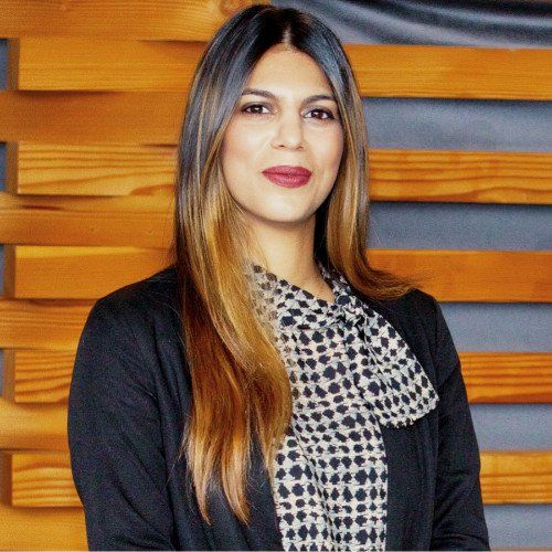 A woman wearing a scarf and a black jacket is standing in front of a wooden wall.