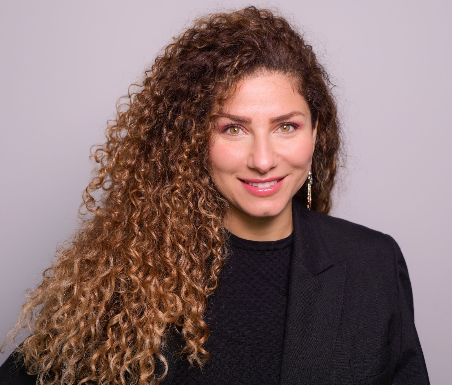 A woman with curly hair is wearing a black jacket and smiling.