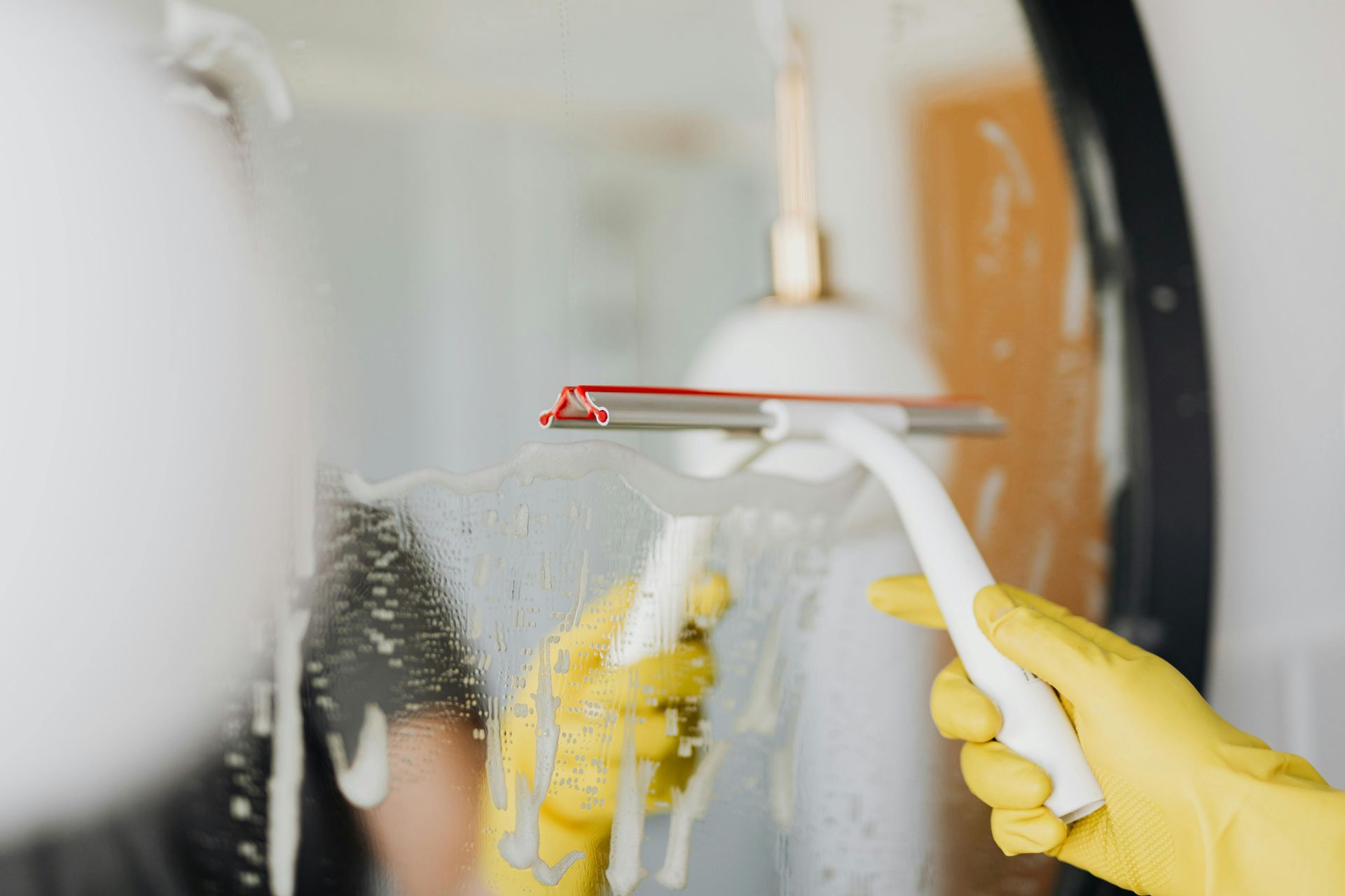 Person in yellow gloves using a squeegee to clean a soapy, round bathroom mirror.