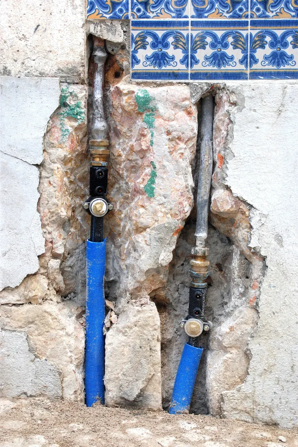 Pipes emerging from a damaged wall, blue covering, ornate blue and white tile at top.