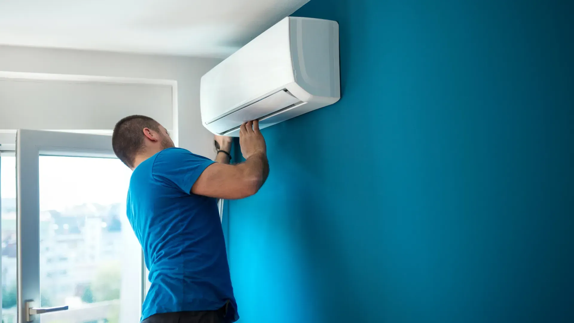 Man in blue shirt installing a white air conditioning unit on a teal wall.