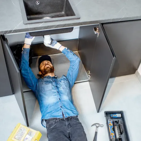 Plumber in blue denim shirt and gloves working under kitchen sink. Tools nearby.