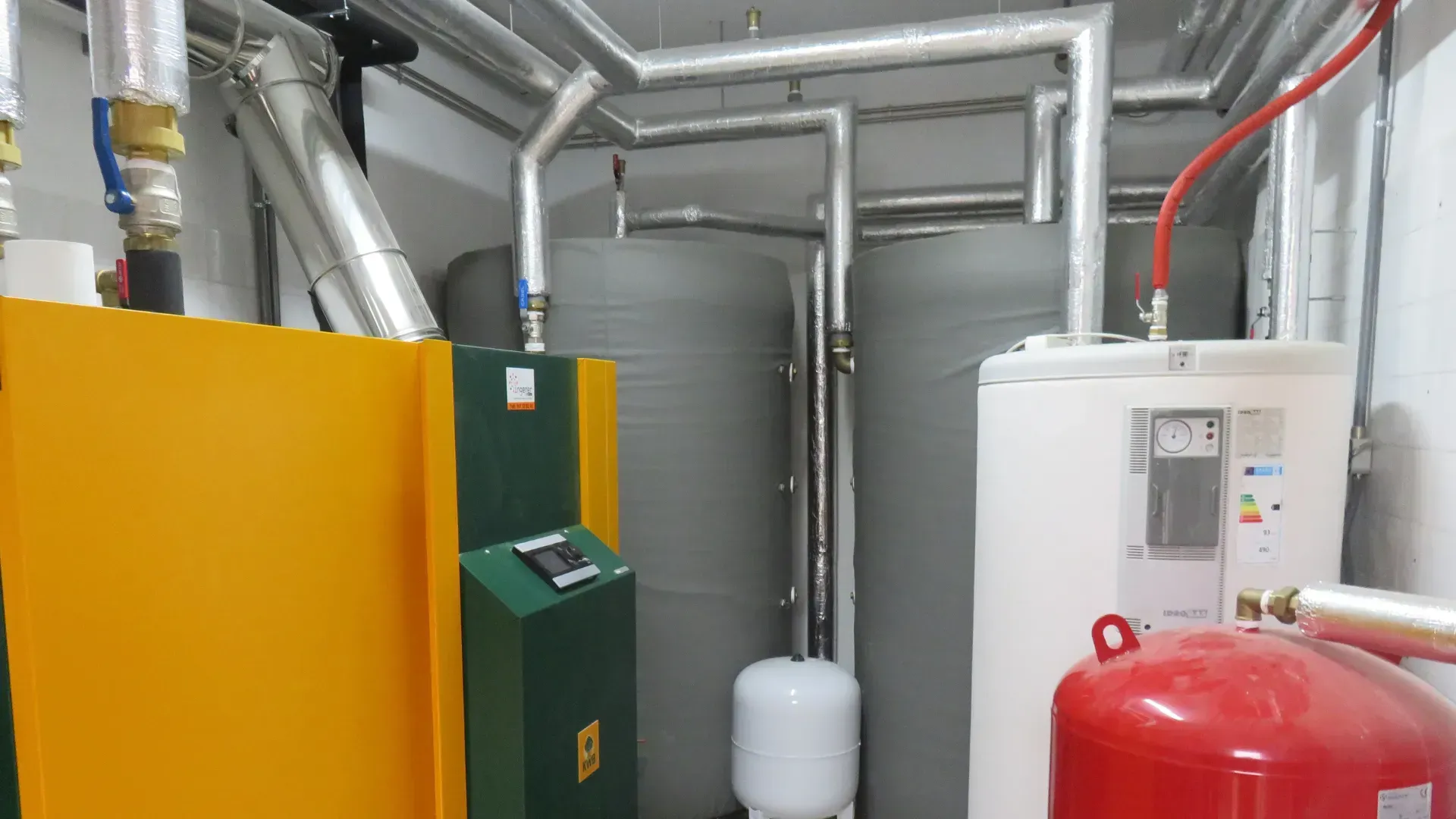 Yellow and green boiler next to gray storage tanks and a white water heater in an equipment room.