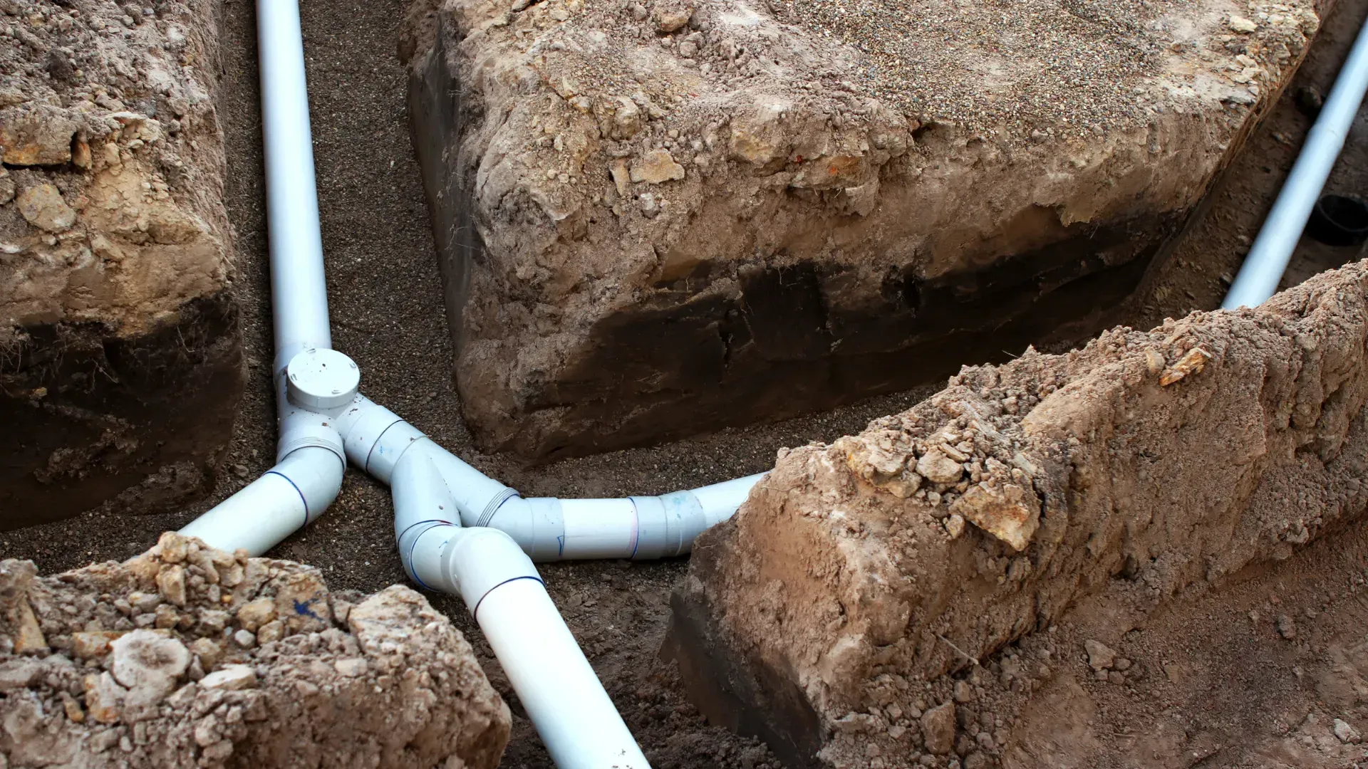 White PVC pipes in trenches, connecting with Y-shaped fittings in dirt.