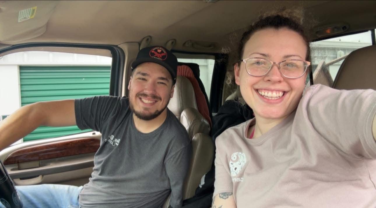 A smiling couple in the front seats of a vehicle, parked in front of green storage units.