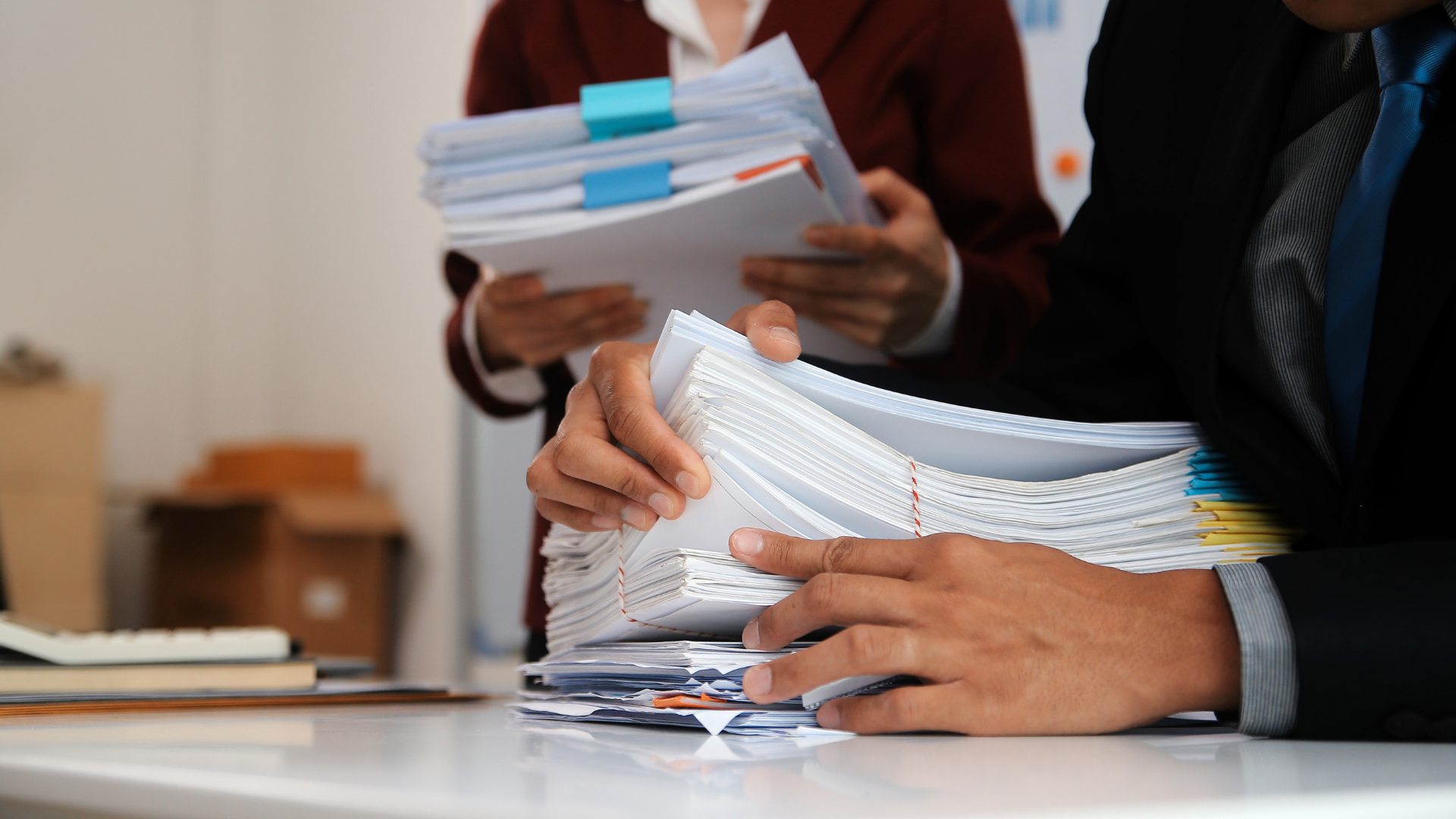 Two people in suits holding large stacks of paperwork, likely in an office.