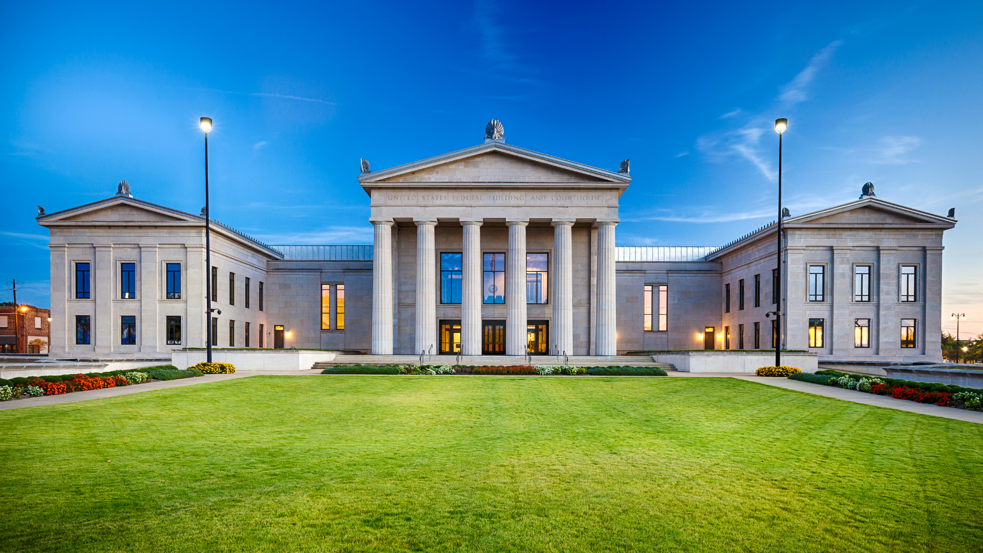 Classical style building with columns, central entrance, flanking wings; green lawn in front, blue sky.