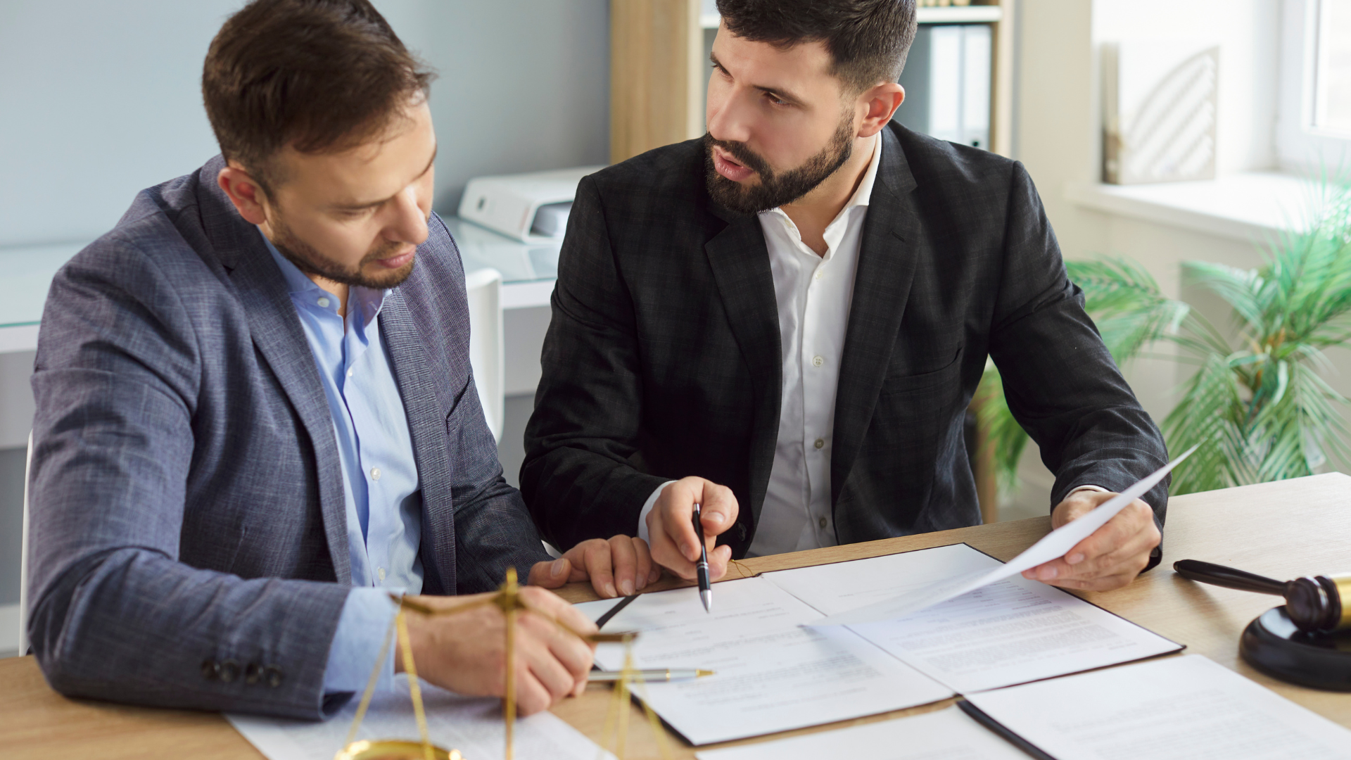 Two men in suits review documents at a desk, with a gavel and scales in the foreground.