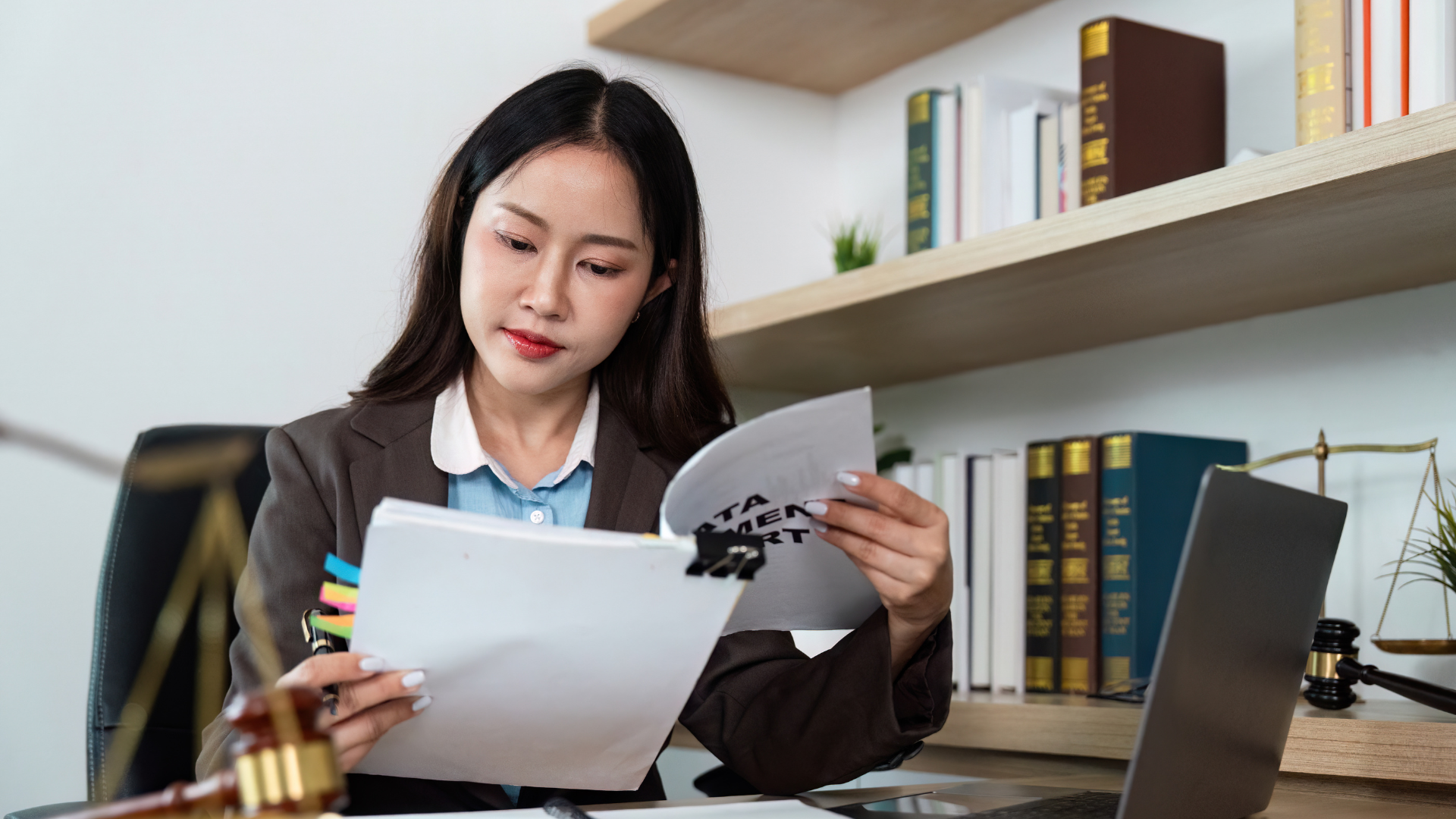 Asian woman in a blazer reviews documents at a desk, with a laptop, books, and a gavel nearby.