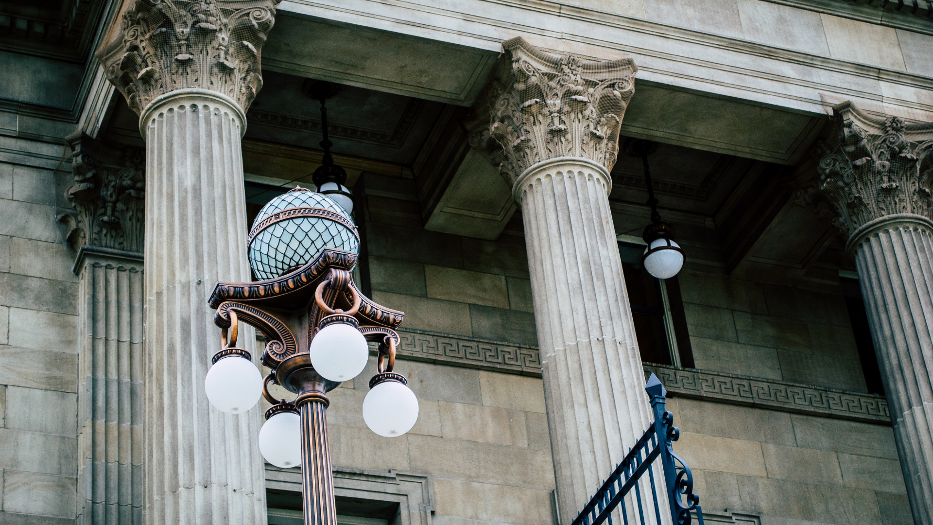 Ornate lamp post with multiple white orbs in front of a stone building with Corinthian columns.