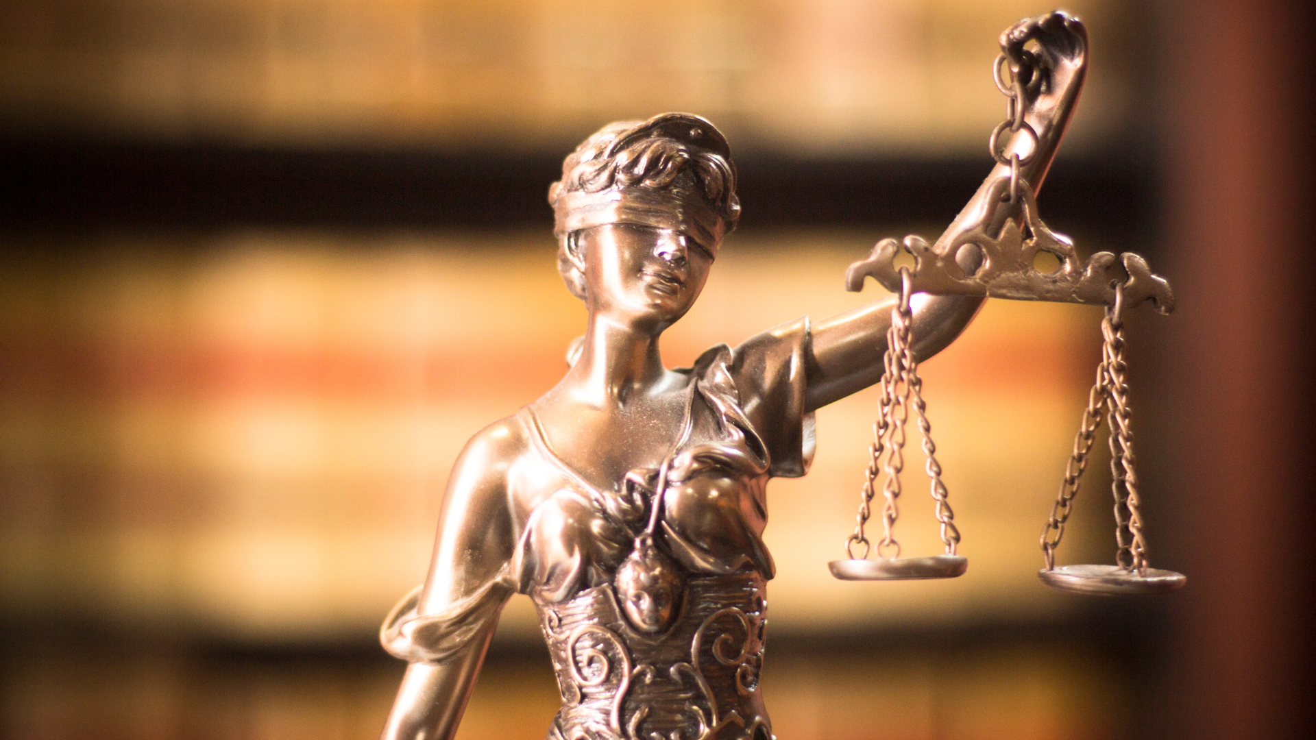 Statue of Lady Justice, blindfolded, holding scales, against a blurred background of bookshelves.