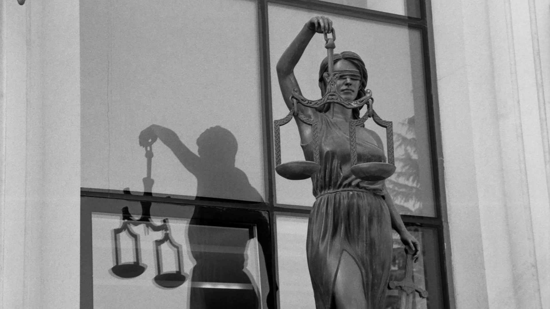 Statue of Lady Justice holding scales, outside a building with reflective windows.