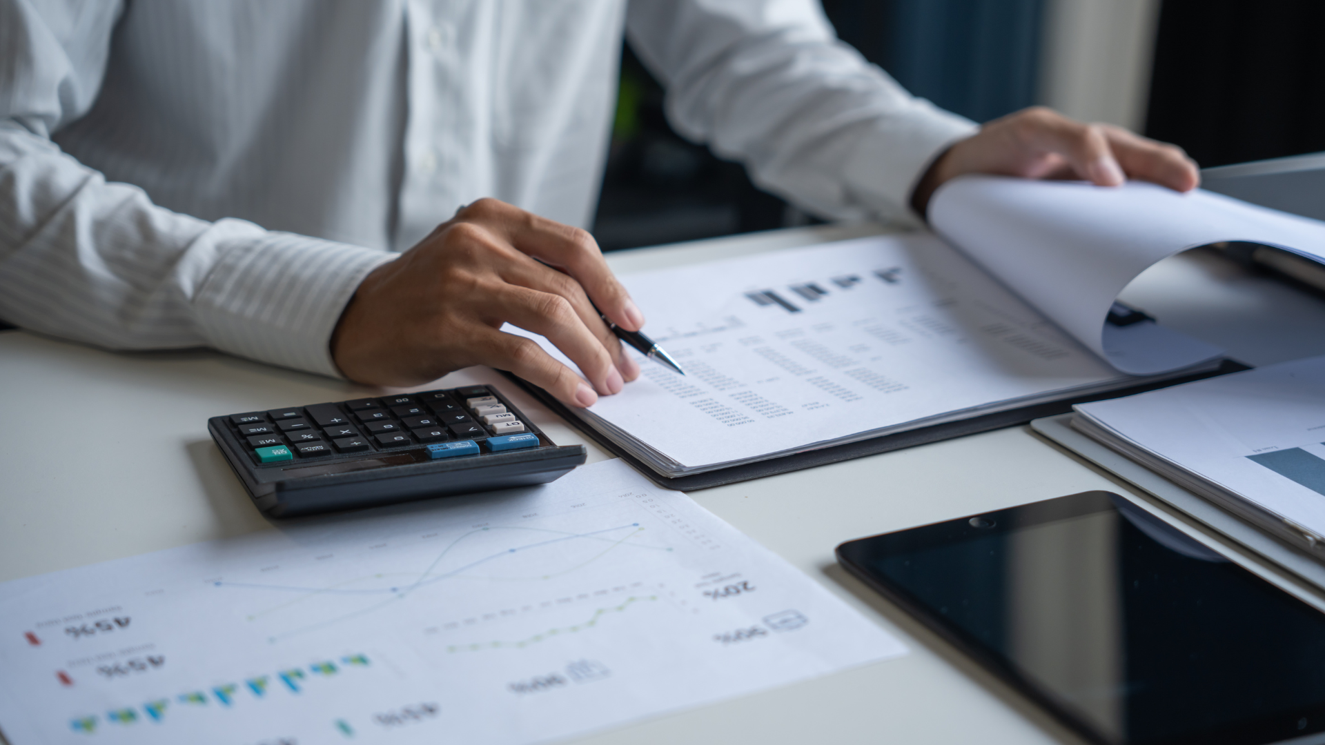 Person in white shirt reviewing financial documents with a pen, calculator, and tablet on a desk.