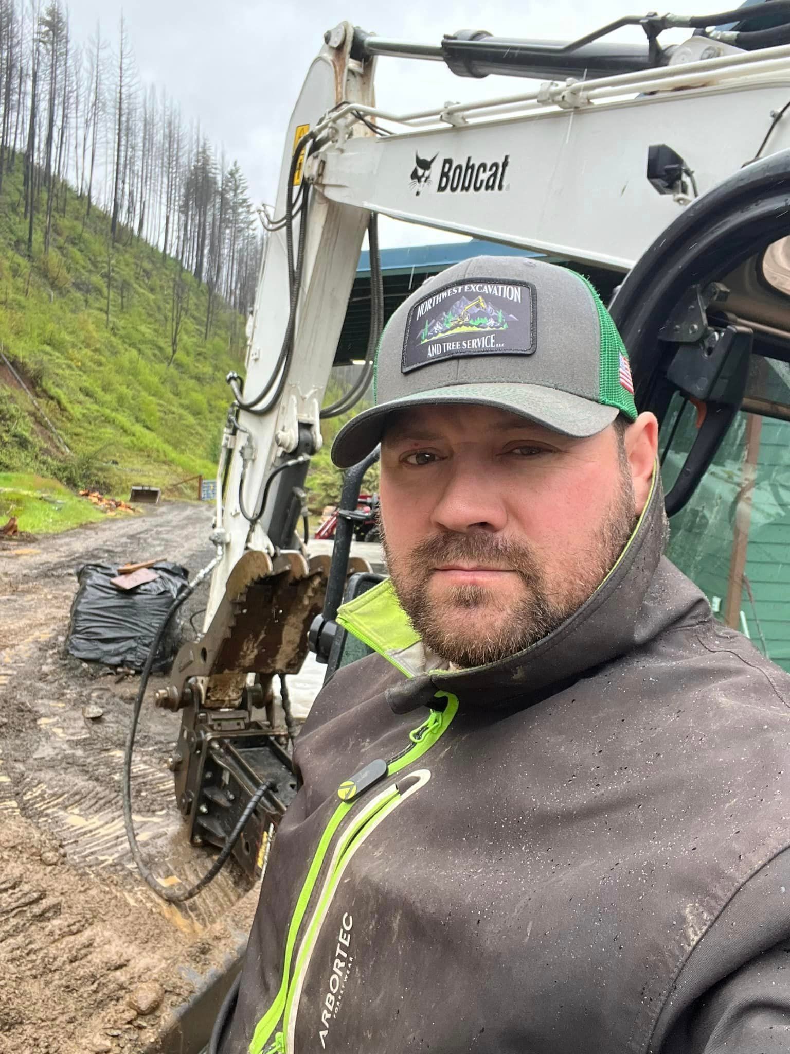 A man wearing a hat and a jacket is standing in front of a bobcat excavator.