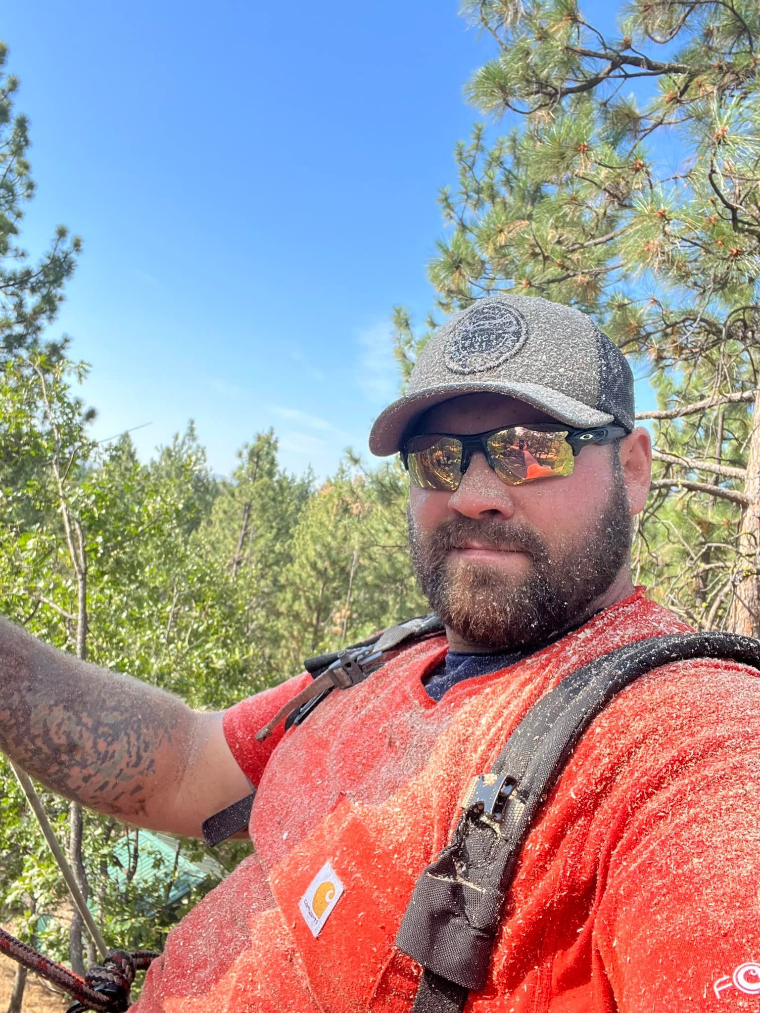 A man wearing sunglasses and a hat is taking a selfie in the woods.