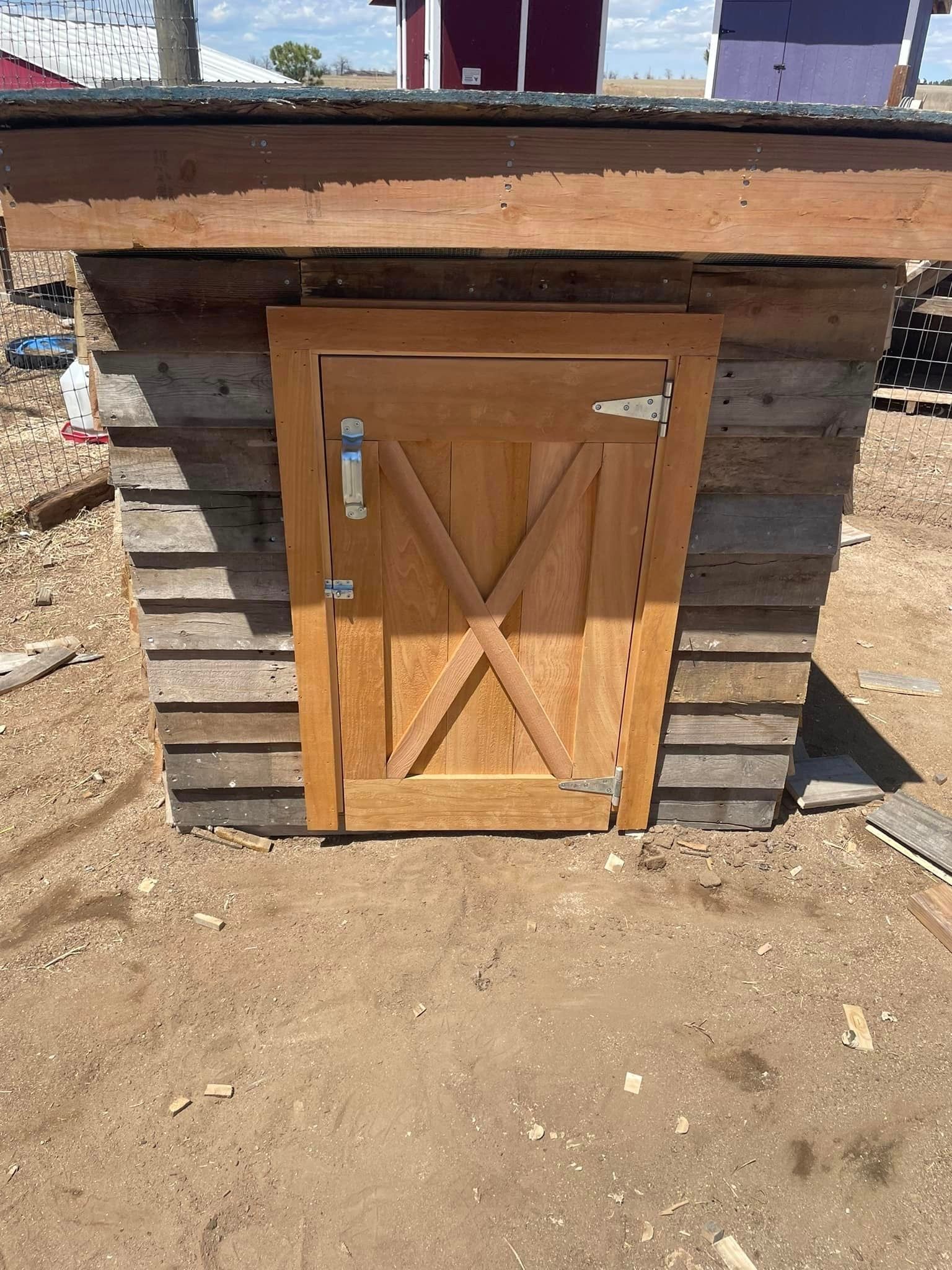 A small wooden shed with a wooden door in the middle of a dirt field.