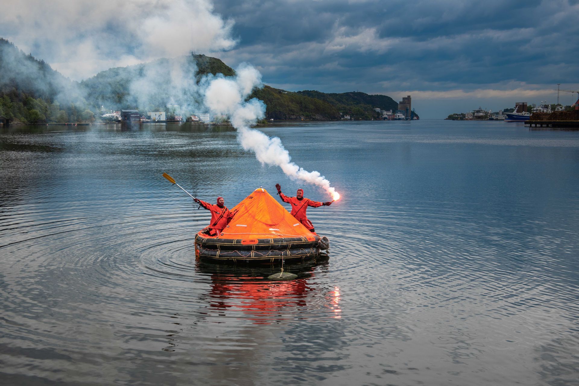 To personer i oransje overlevelsesdrakter på en oppblåsbar flåte i vannet, med fakler i hånden. Røyk stiger opp i en havneomgivelse.
