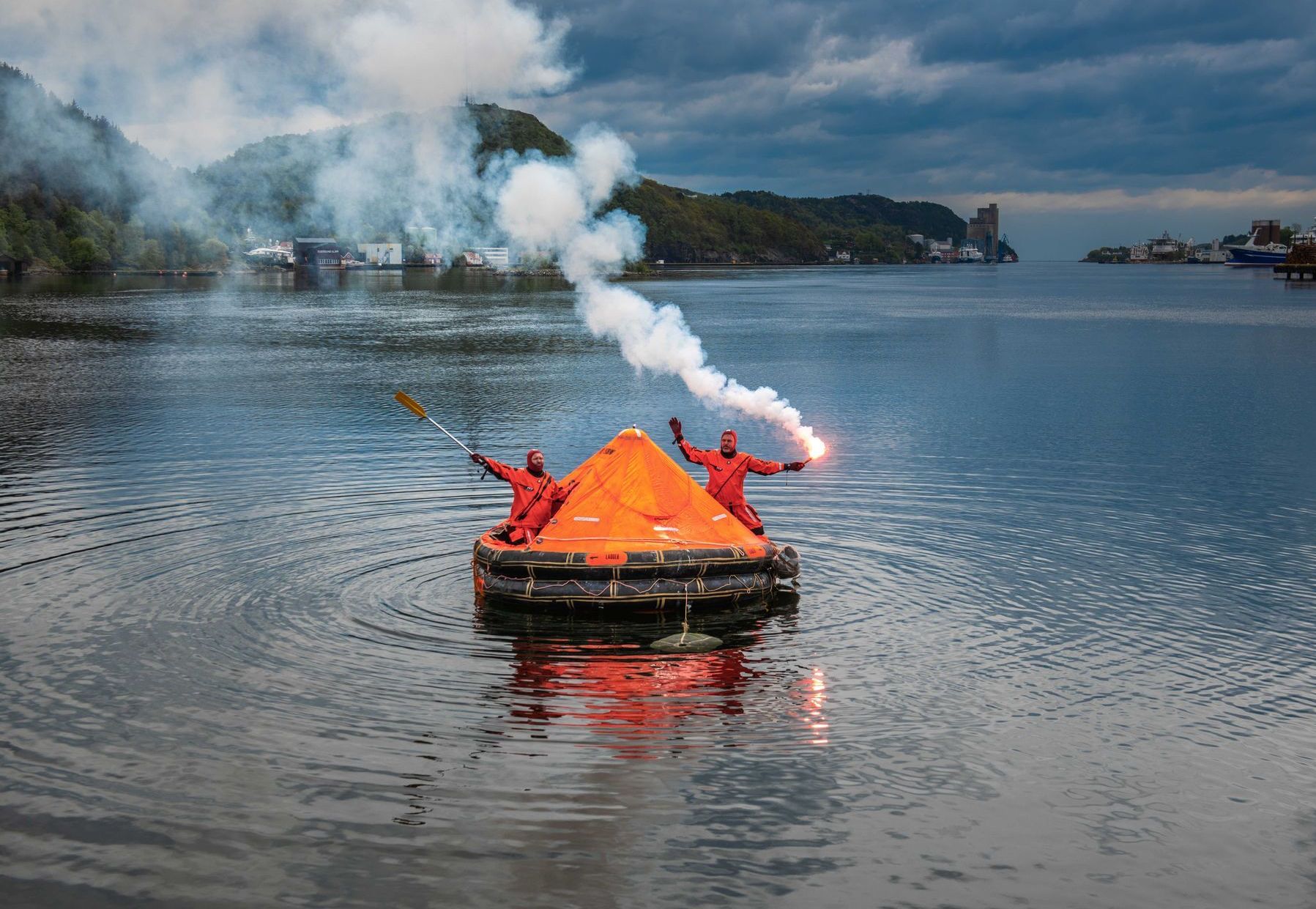 To personer i røde drakter på en oransje redningsflåte i vannet, med fakler som avgir hvit røyk. Et fjellandskap er i bakgrunnen.
