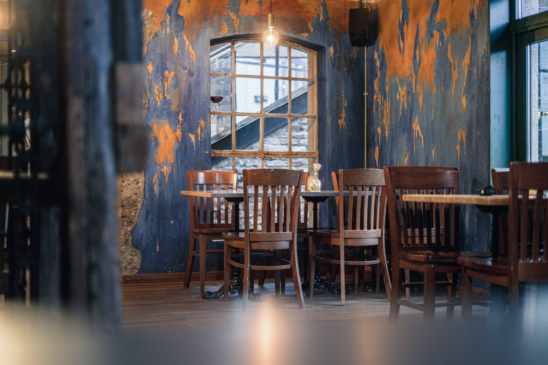 Tomt restaurantinteriør med trestoler og -bord mot avskallende, fargerikt malte vegger, vindu i bakgrunnen.