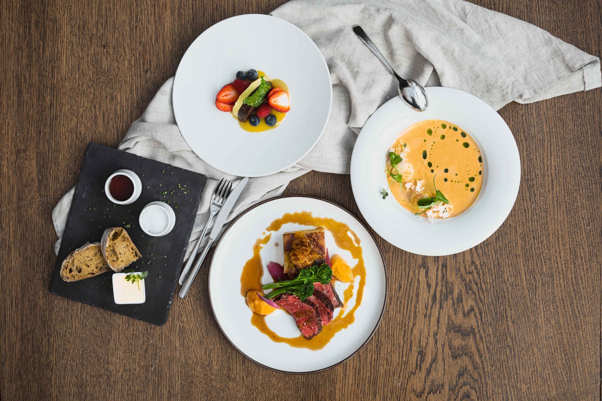 Oversiktsbilde av et trebord med fire tallerkener med mat, inkludert frukt, suppe og biff. En serviett og bestikk er også til stede.