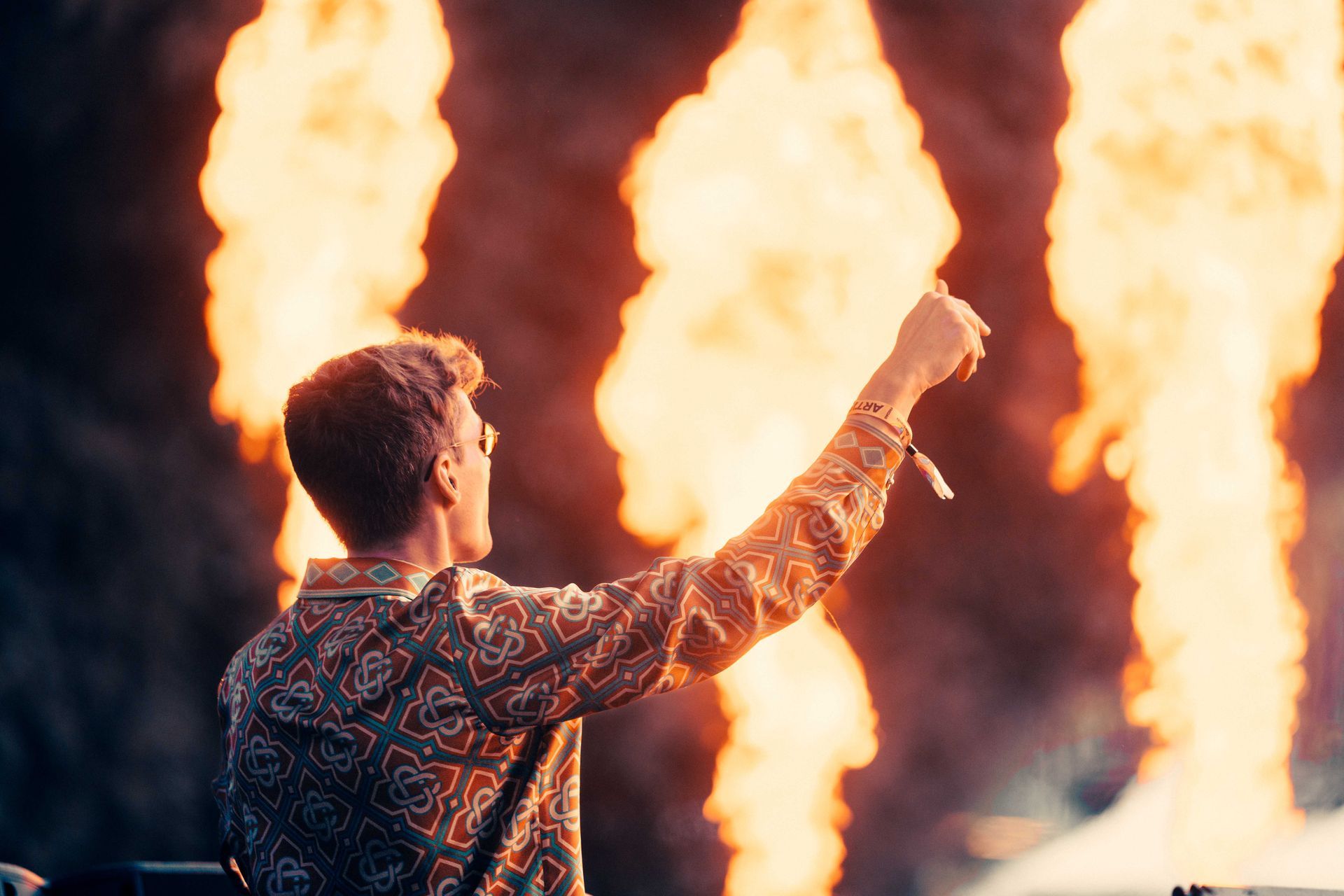 Mann med hevet arm på scenen med flammer i bakgrunnen. Han har på seg en mønstret skjorte og armbånd, sett bakfra.