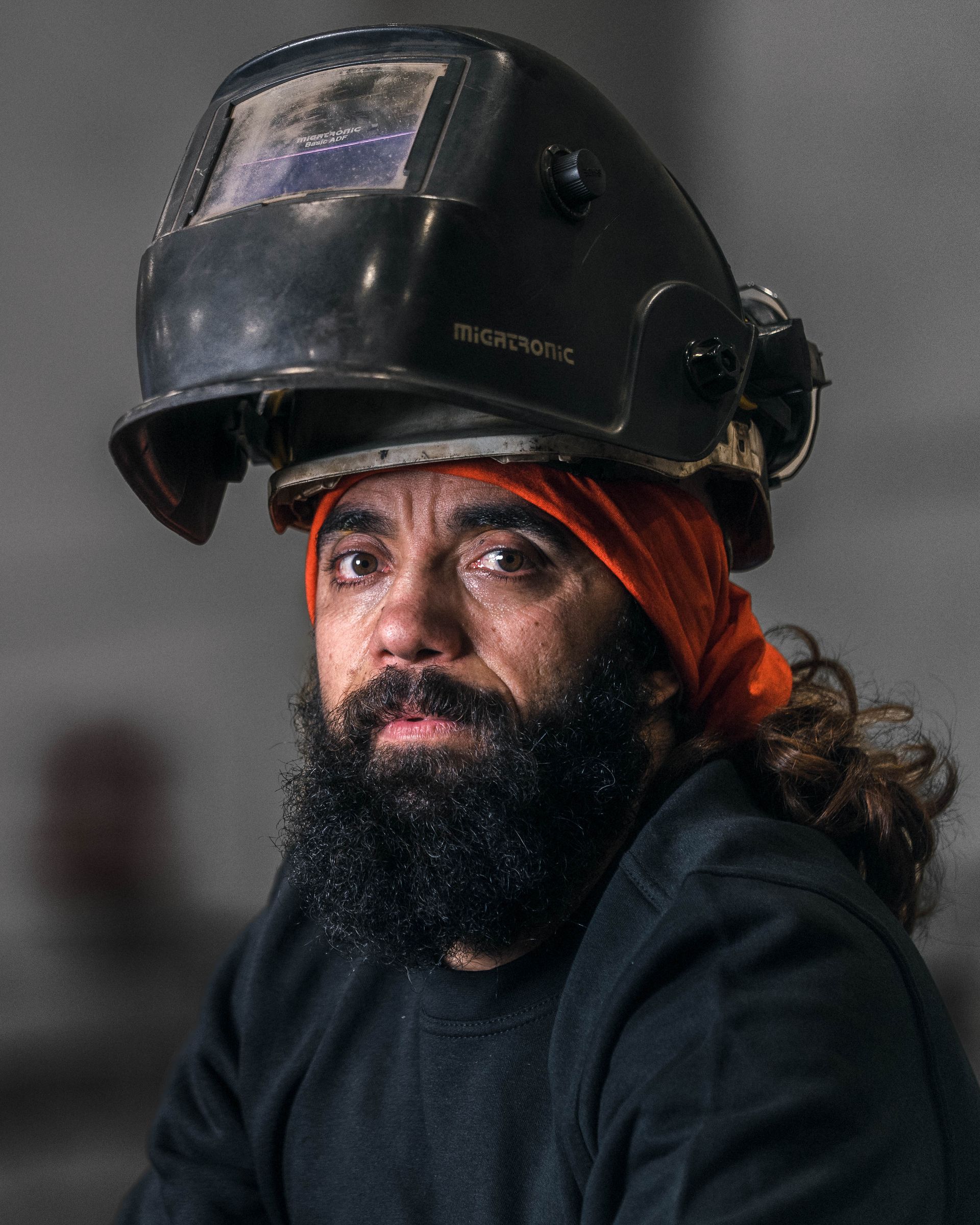 Mann med heldekkende skjegg iført sveisehjelm og oransje hodeskjerf, ser inn i kameraet.