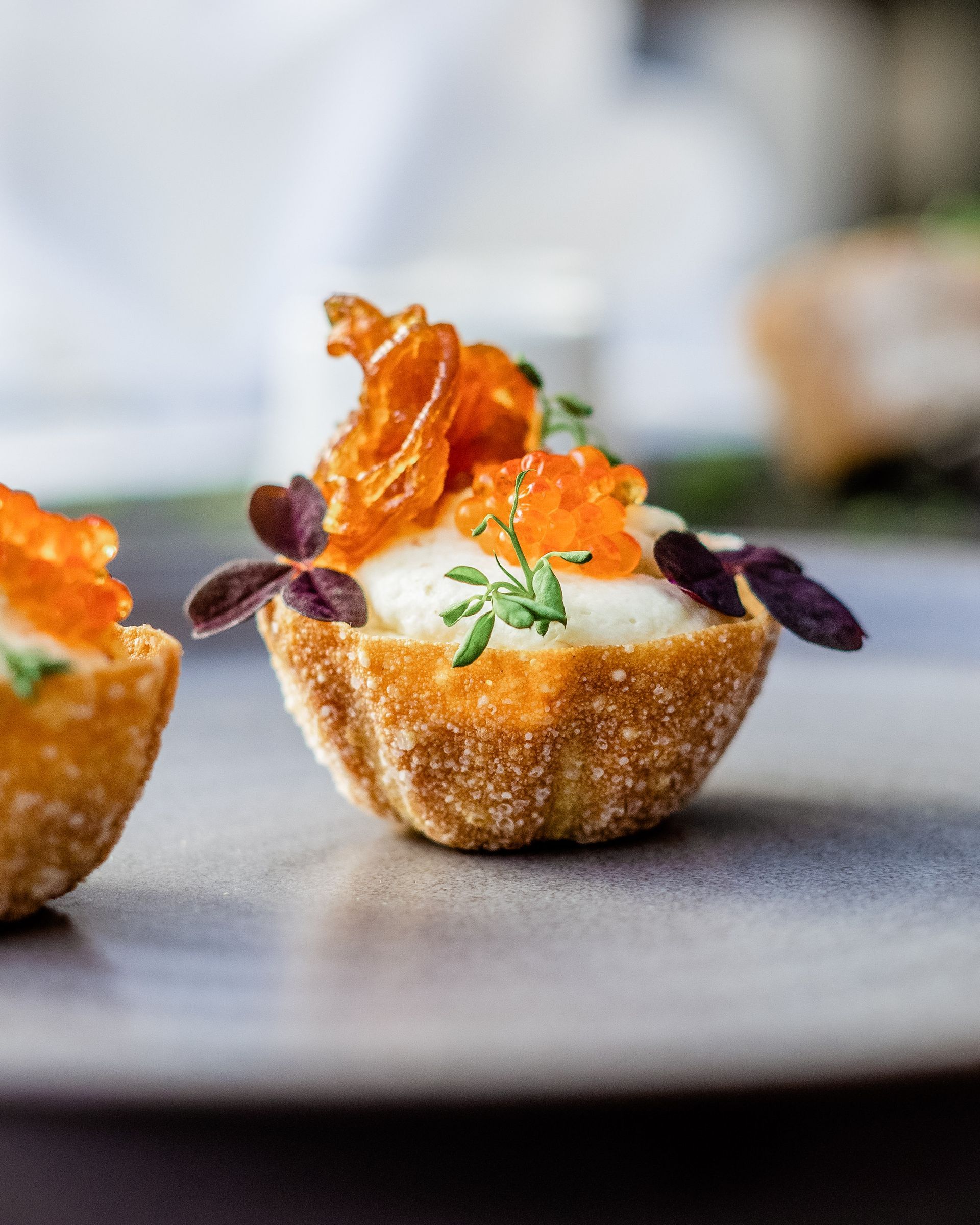 Nærbilde av en enkelt, gyllenbrun tartlett fylt med en hvit mousse, toppet med appelsinrogn og chips, og garnert med lilla blader på en grå tallerken.