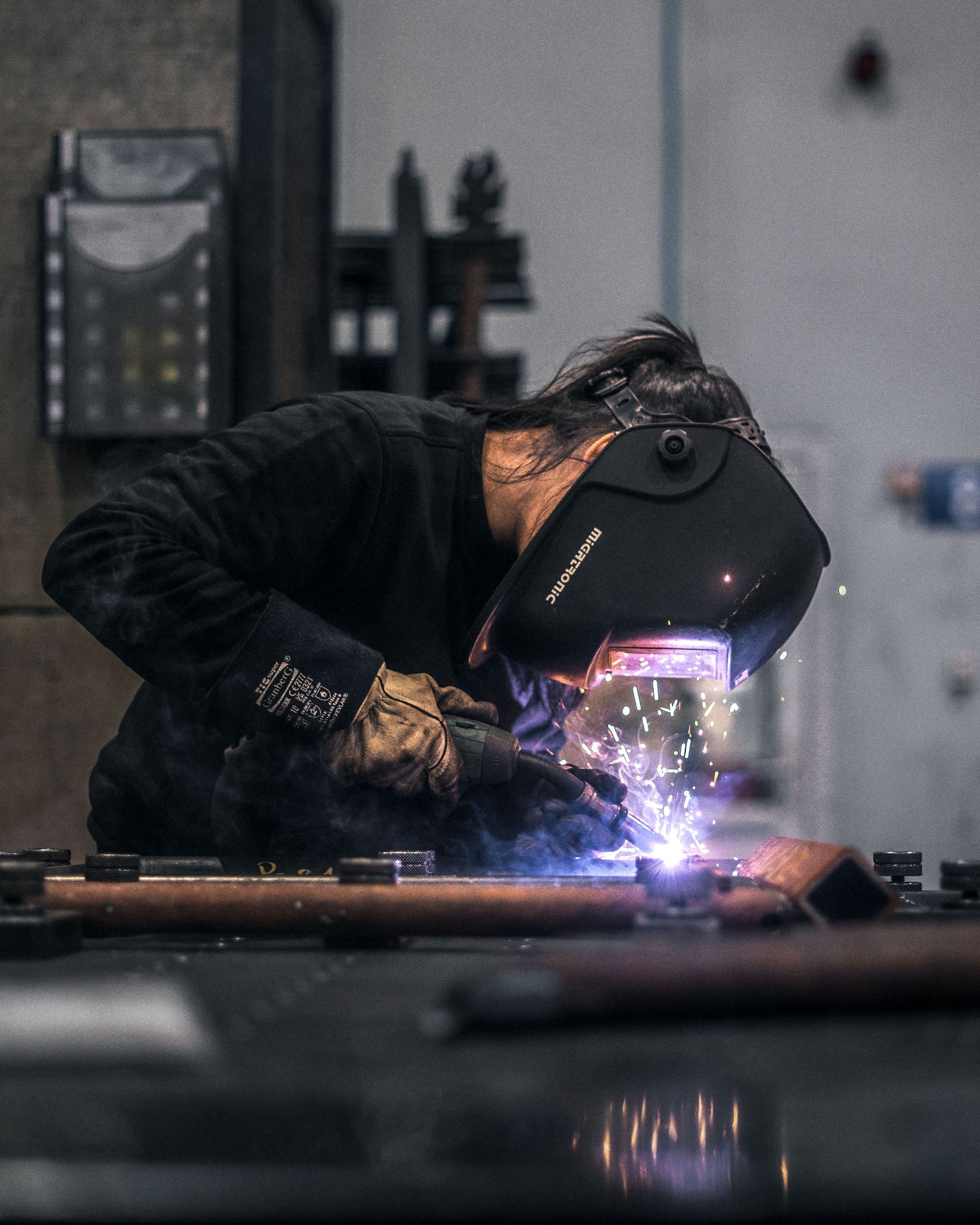 En person som sveiser metall, iført beskyttelsesmaske og hansker, gnister flyr, innendørs.