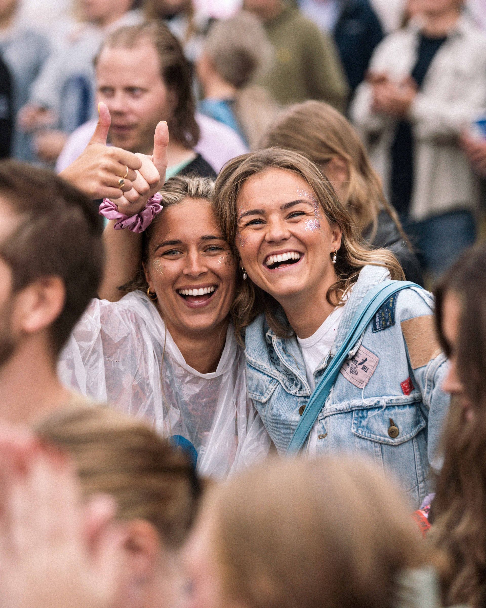 To smilende unge kvinner med glitter i ansiktene omfavner hverandre i en folkemengde og koser seg med et utendørsarrangement. Den ene gir tommelen opp.