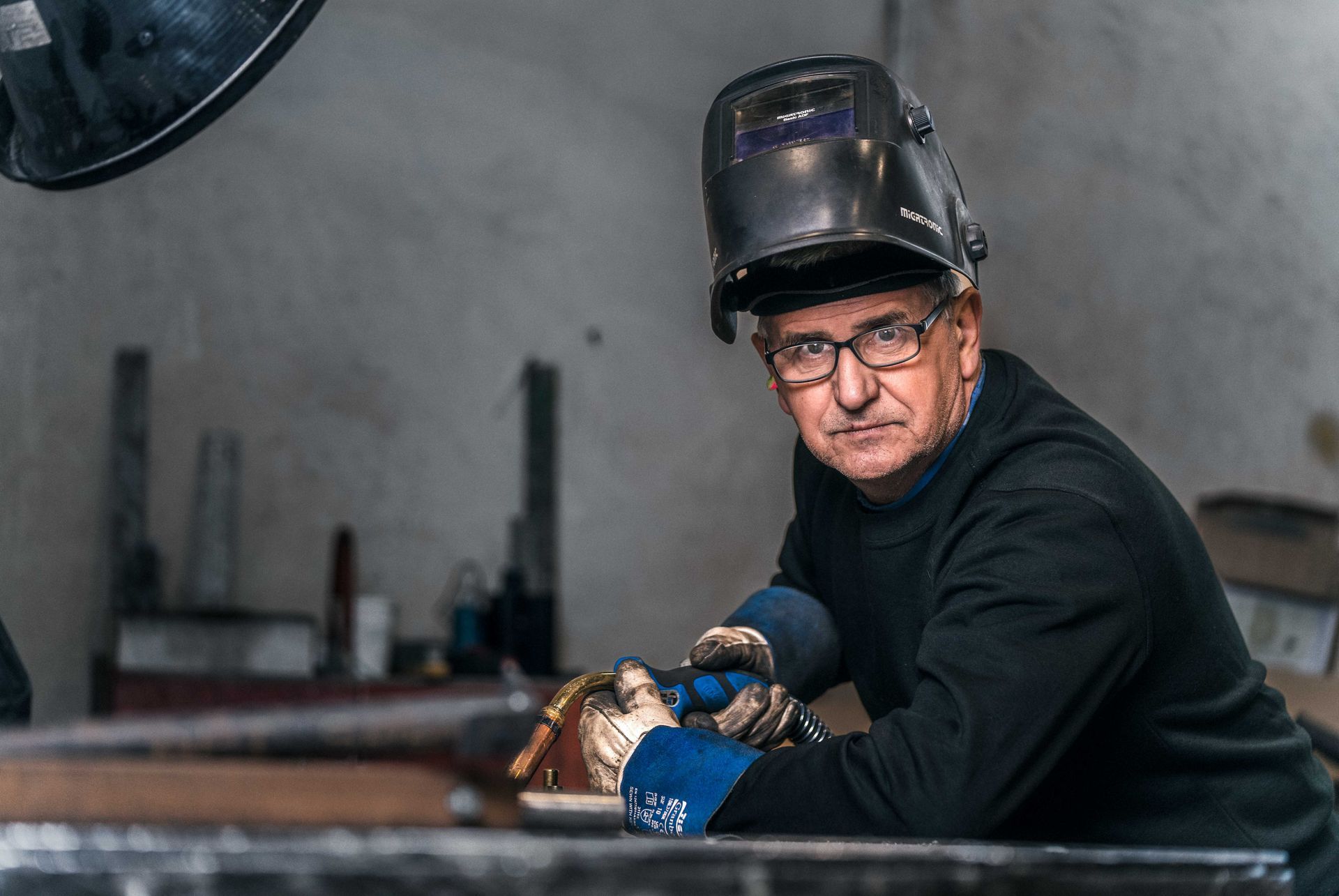 Mann i sveisehjelm og hansker lener seg på en metalloverflate i et verksted. Han har på seg briller og en svart skjorte.