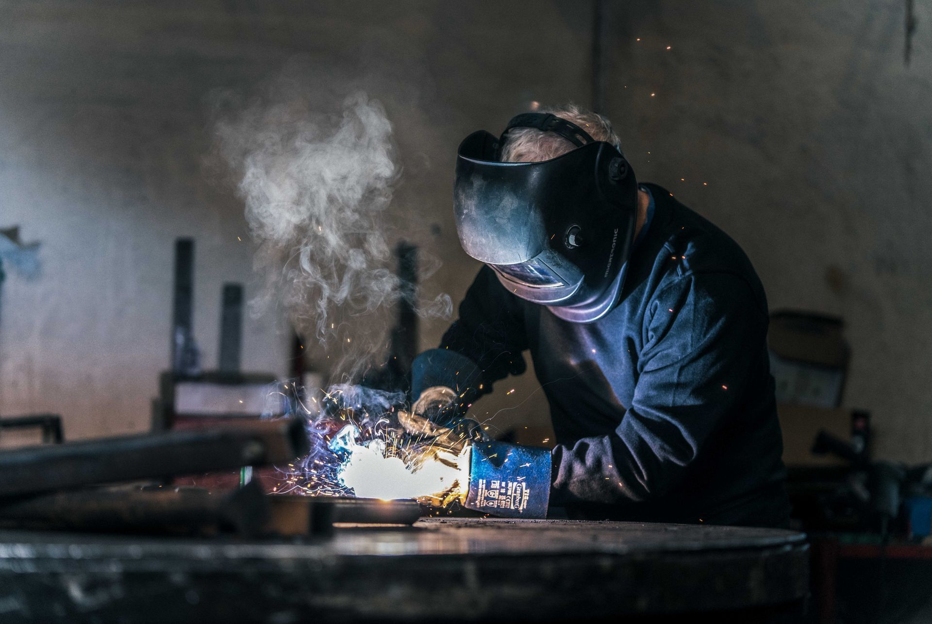 Sveiser i beskyttelsesutstyr arbeider med metall og lager gnister og røyk i et svakt opplyst verksted.