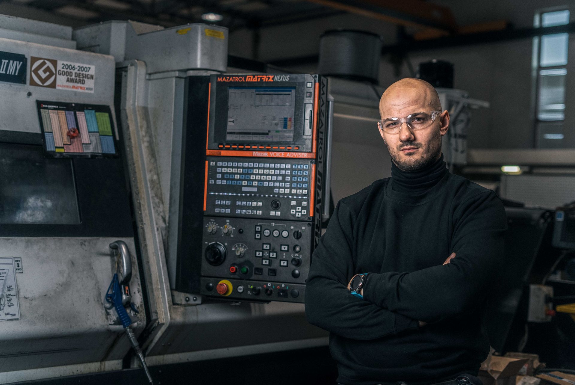 En mann med kryssede armer iført vernebriller står foran en CNC-maskin i et verksted.