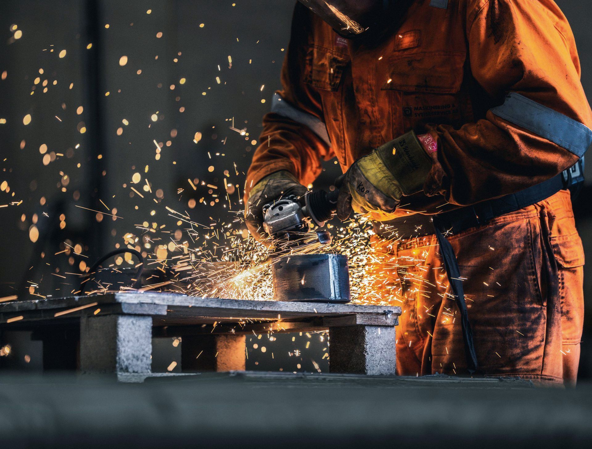 En arbeider i sikkerhetsutstyr sliper metall og lager gnister i et svakt opplyst verksted.