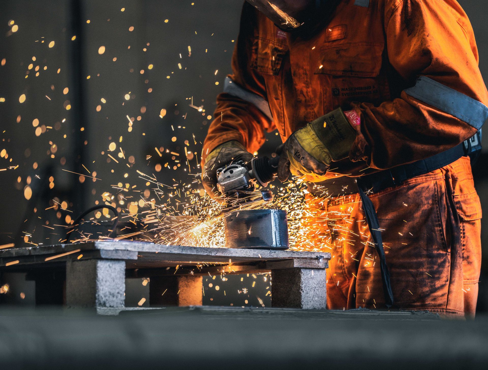 En person i oransje beskyttelsesutstyr bruker en kvern og lager gnister over en metallgjenstand på en trepall.