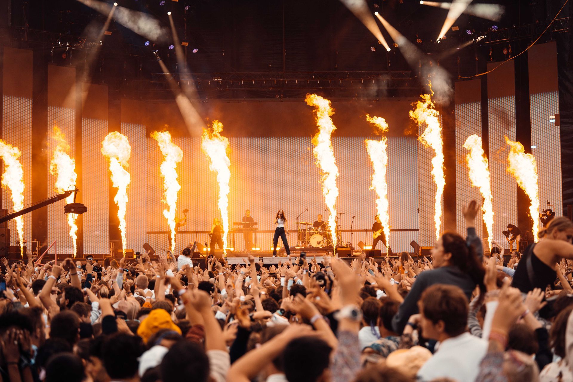 En konsertscene med en sanger og et band som opptrer foran en stor folkemengde. Flammer bryter ut fra scenen.