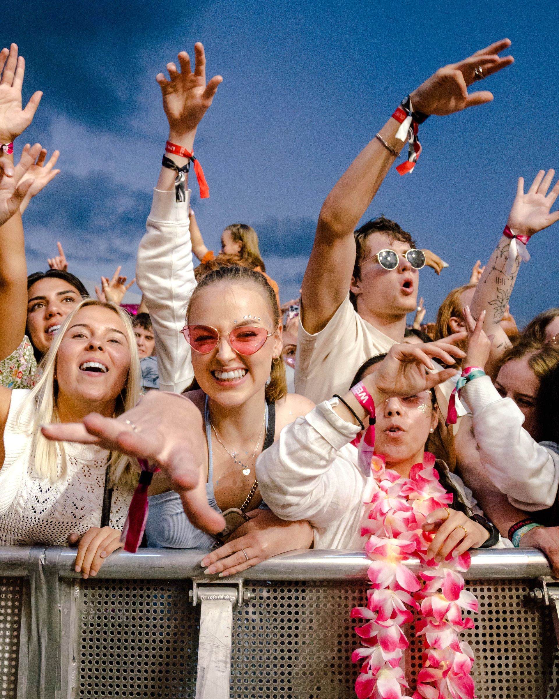 Folk på en musikkfestival, jubler og løfter armene foran en metallbarriere. Noen bruker armbånd og solbriller.