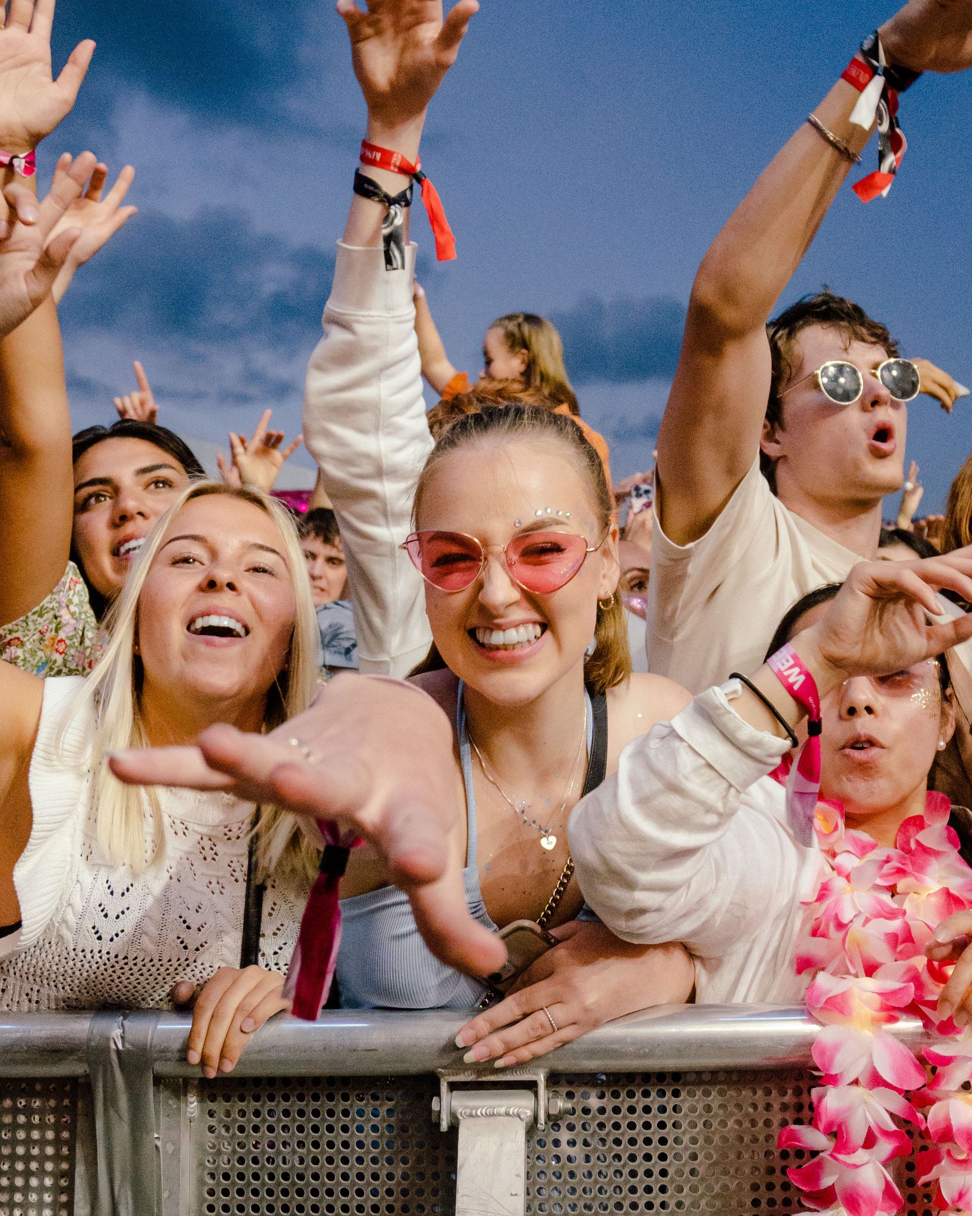 Folk på en musikkfestival med armene hevet, inkludert en kvinne med hjerteformede briller, alle smilende og har det gøy.