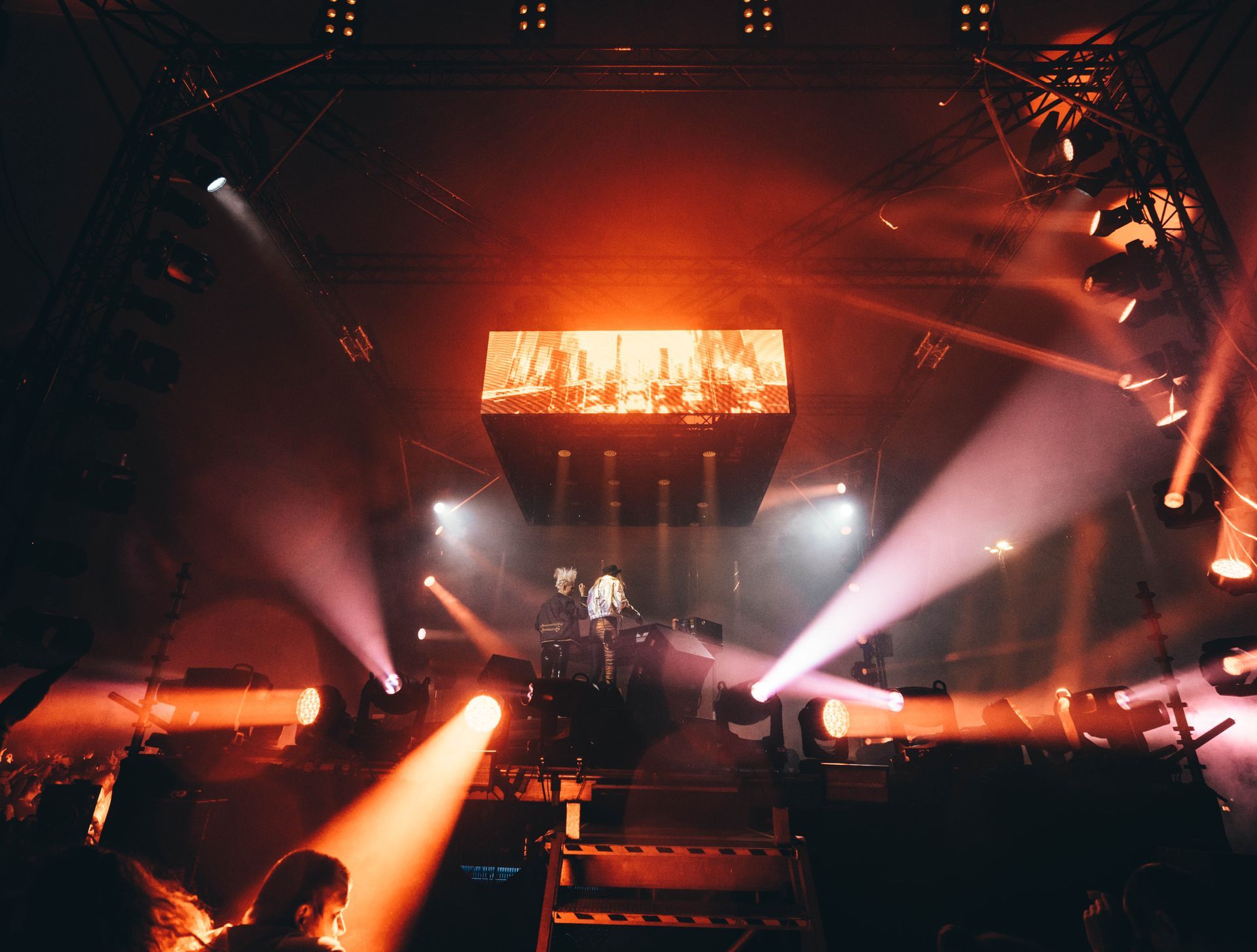 Konsertscene badet i rødt lys, utøvere i silhuett under en stor skjerm, lysstråler som stråler utover.